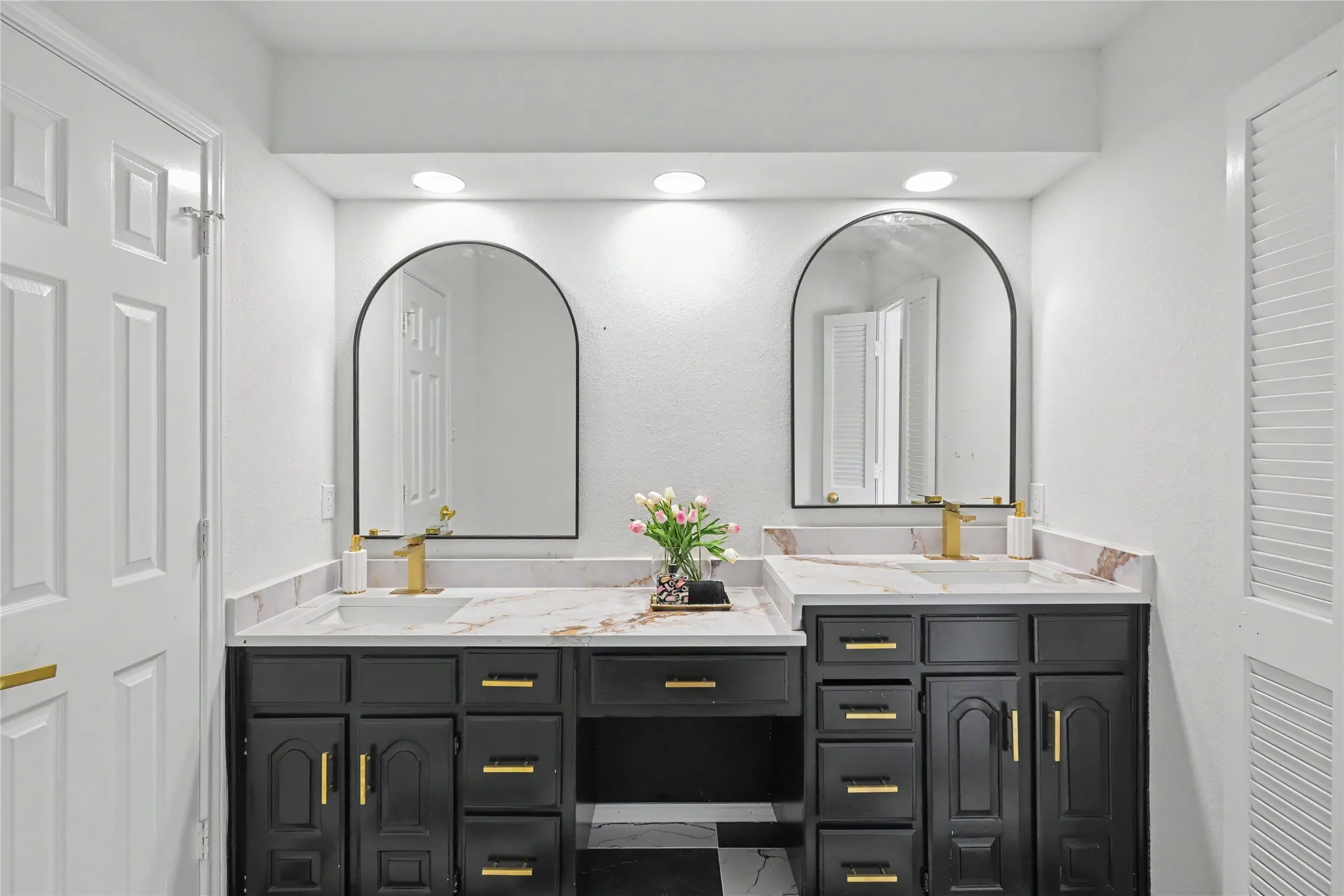 Double vanity with dark-finish cabinetry, gold-tone hardware, stone-composite countertop, arched mirrors, and gold-tone fixtures
