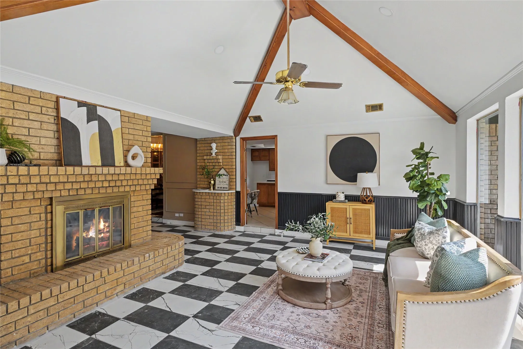 Vaulted ceiling with exposed wood beams, brick fireplace featuring a metal insert, checkerboard tile flooring, and dark wainscoting