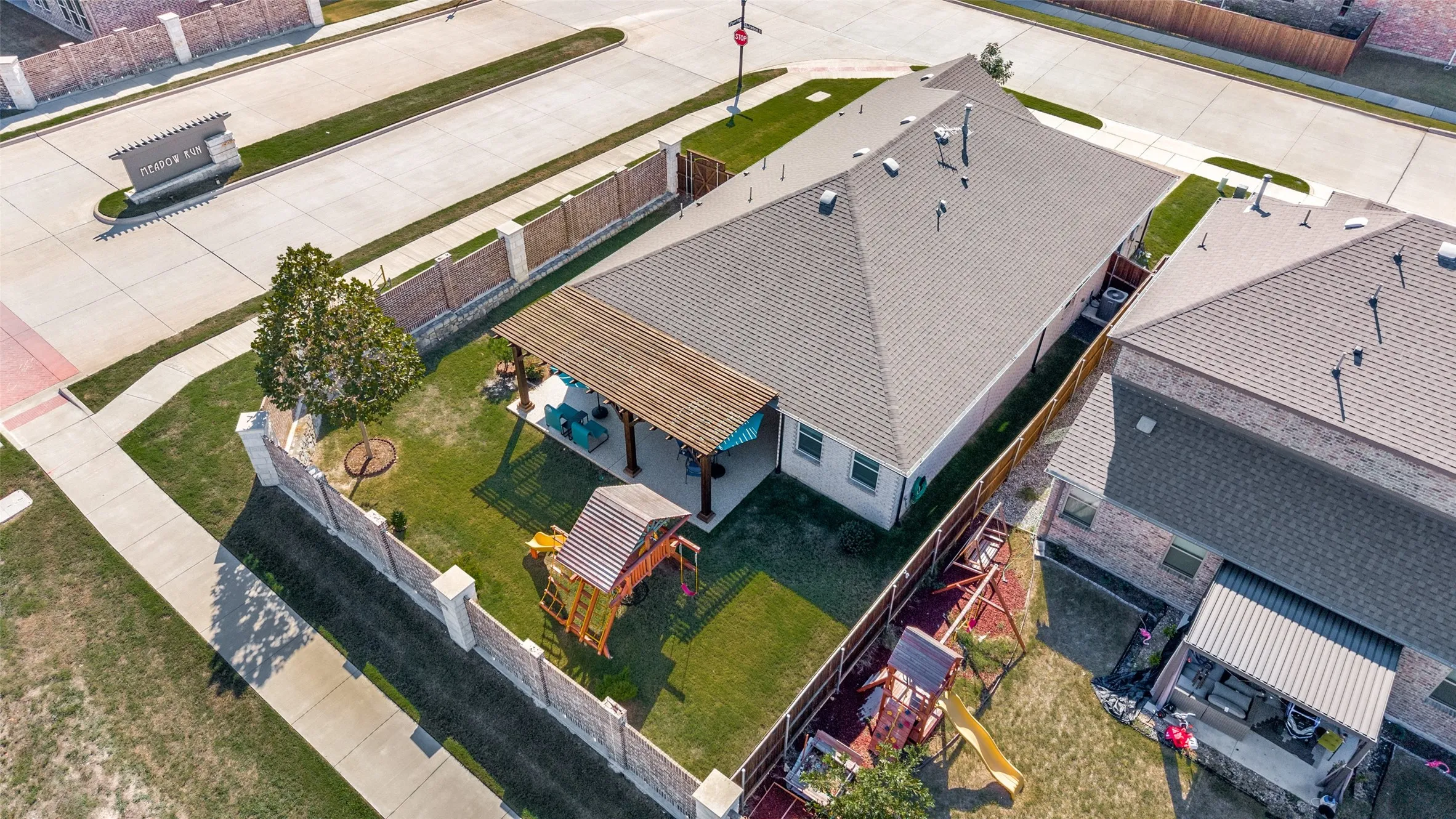 Aerial perspective of a corner lot property featuring a stone and brick perimeter wall, a covered patio with a wooden pergola, and a private lawn area