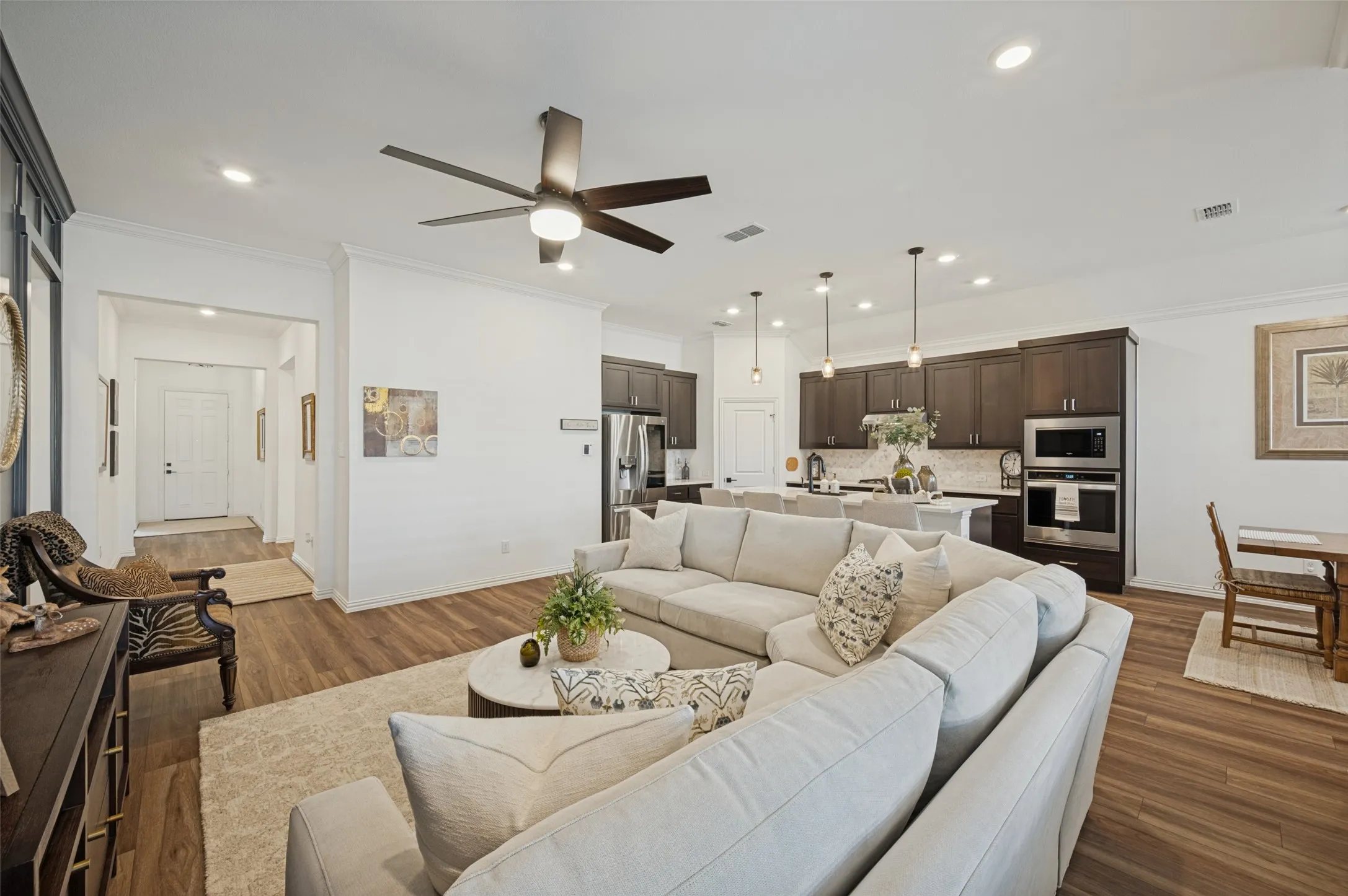 Open-concept interior featuring wood-finish flooring, recessed ceiling lighting, a contemporary multi-blade ceiling fan, and a kitchen equipped with dark cabinetry, stone countertops, and built-in stainless steel appliances