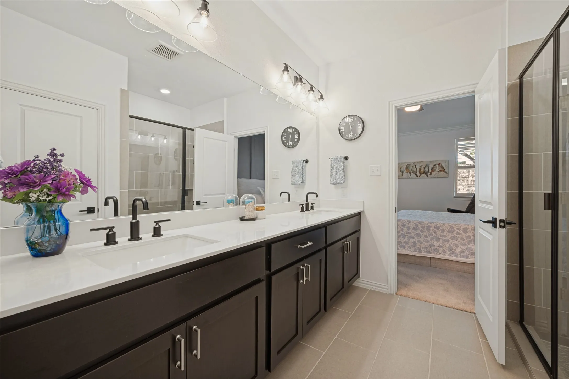 Dual-sink vanity with matte black fixtures, crisp white countertop, expansive wall-to-wall mirror, and glass-enclosed shower with neutral-toned tiling