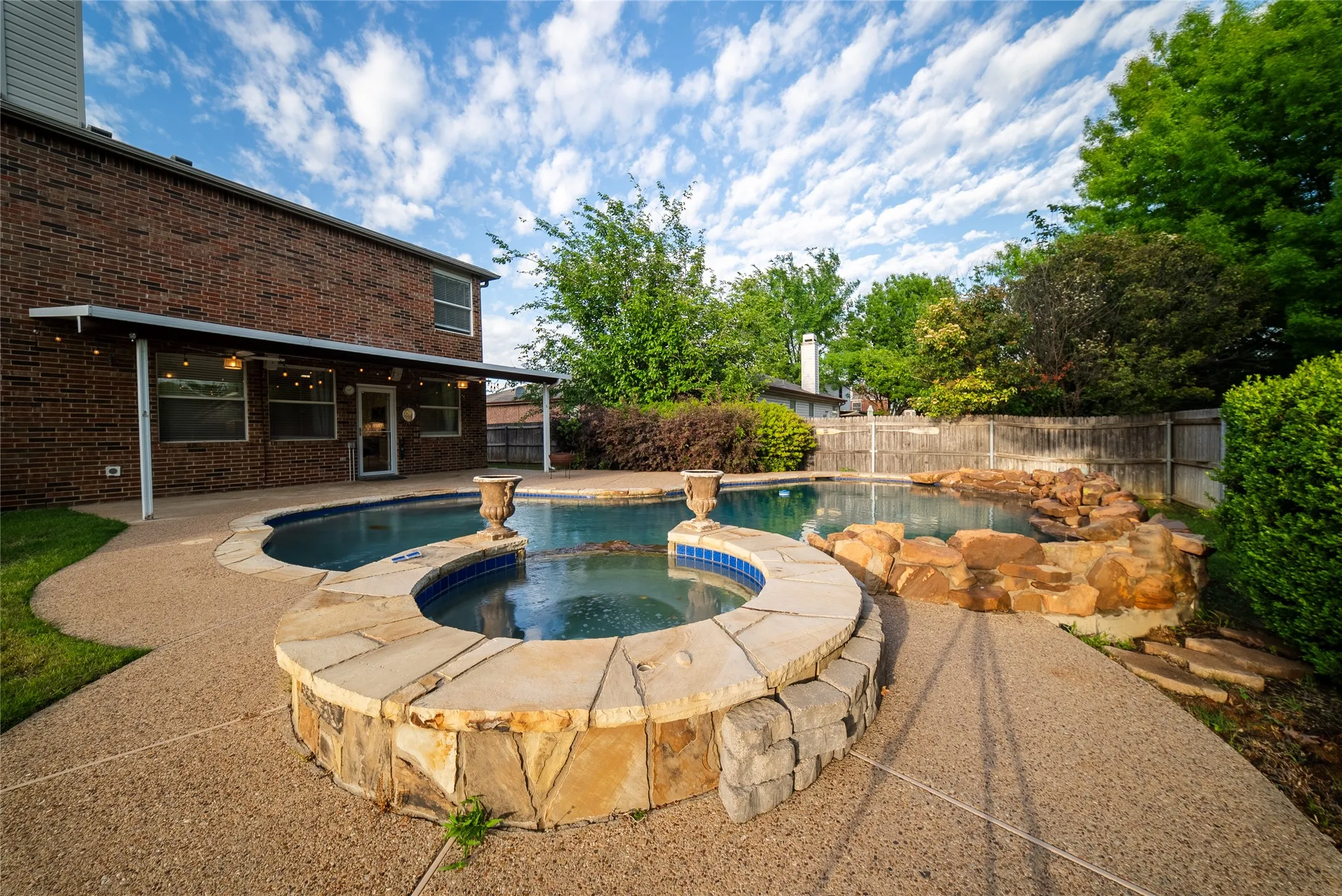 Backyard featuring an inground pool, elevated stone-clad spa, natural rock water feature, and a textured concrete patio