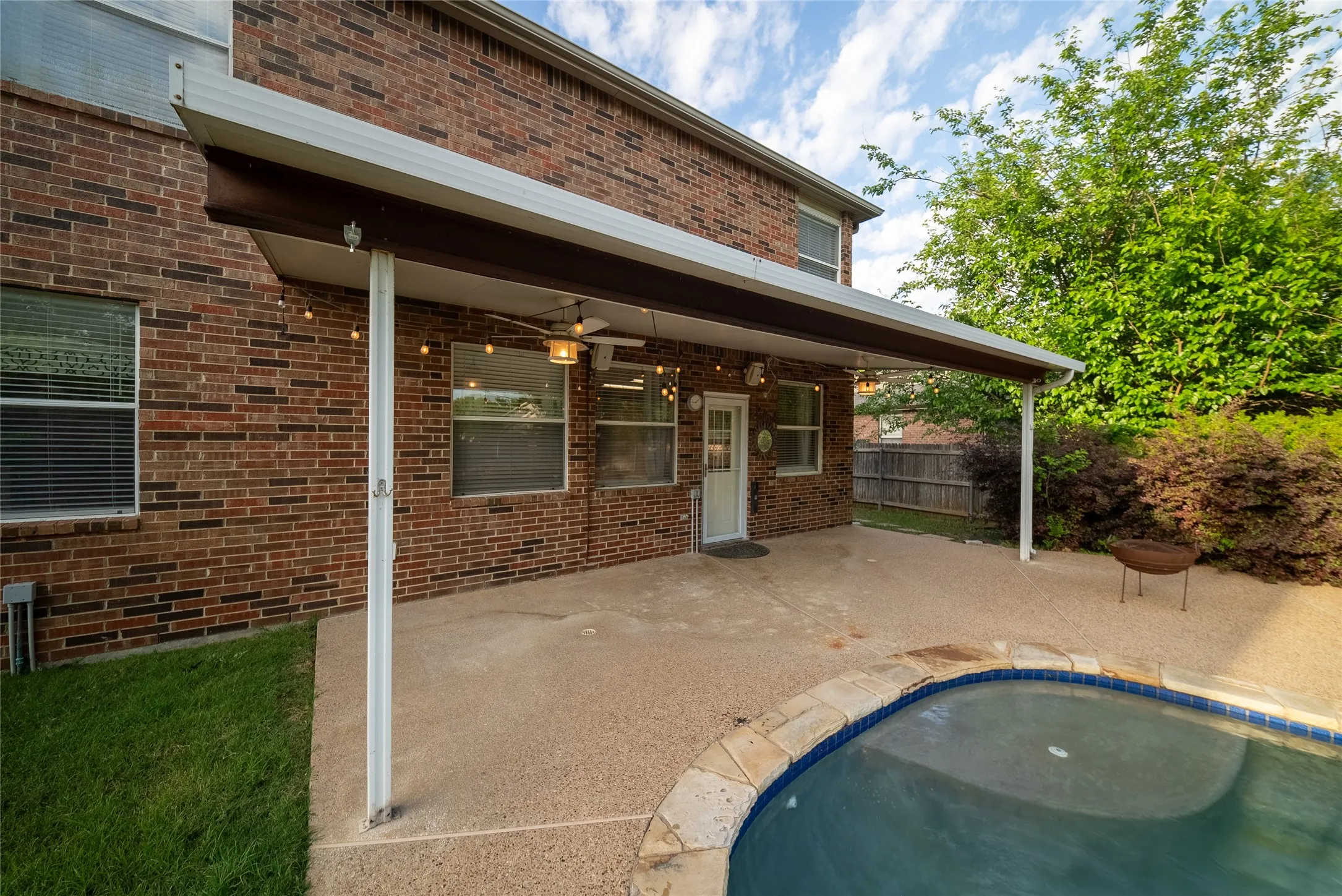 Covered patio with aggregate concrete flooring, brick exterior siding, and an inground swimming pool featuring stone coping