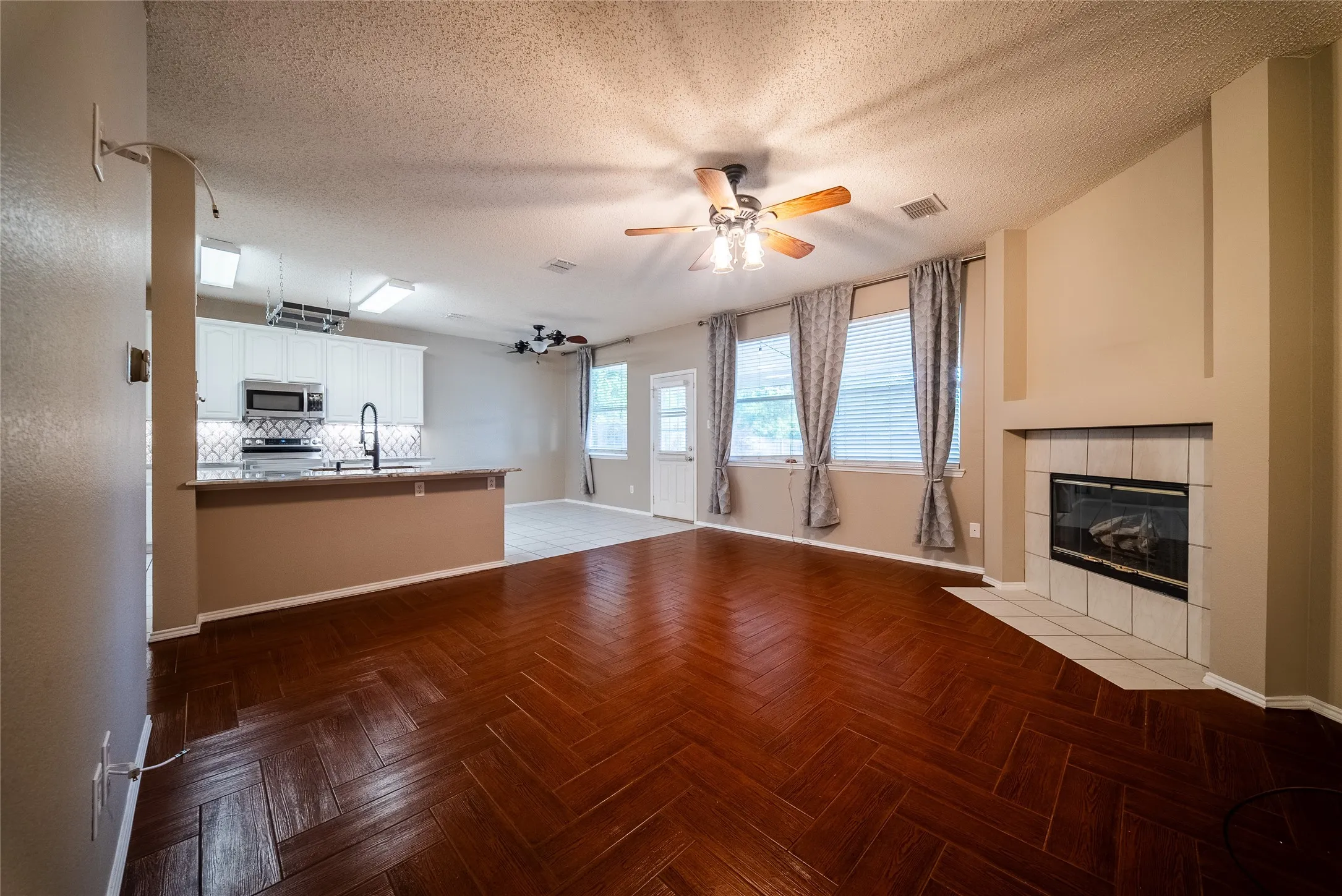 Herringbone-pattern wood-finish flooring, built-in fireplace with tiled surround, white kitchen cabinetry, breakfast bar, and multiple ceiling fans
