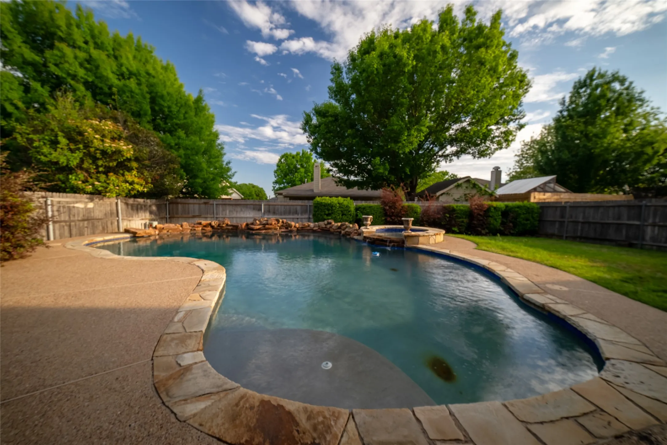 In-ground pool featuring a stone-accented spa, rock water feature, textured concrete decking, and perimeter wood fencing