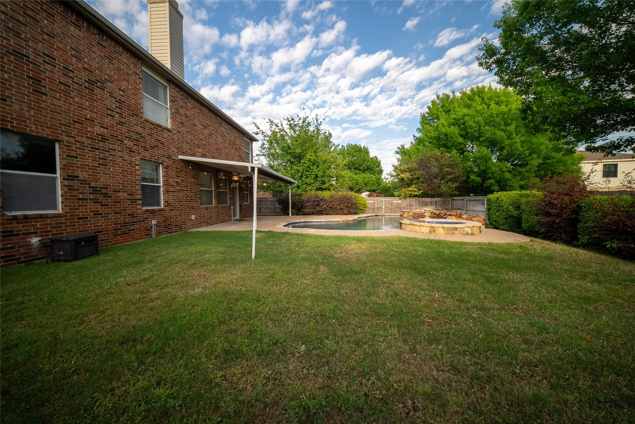 Brick exterior siding featuring a covered patio, swimming pool with a stone-accented spillover spa, and mature landscaping