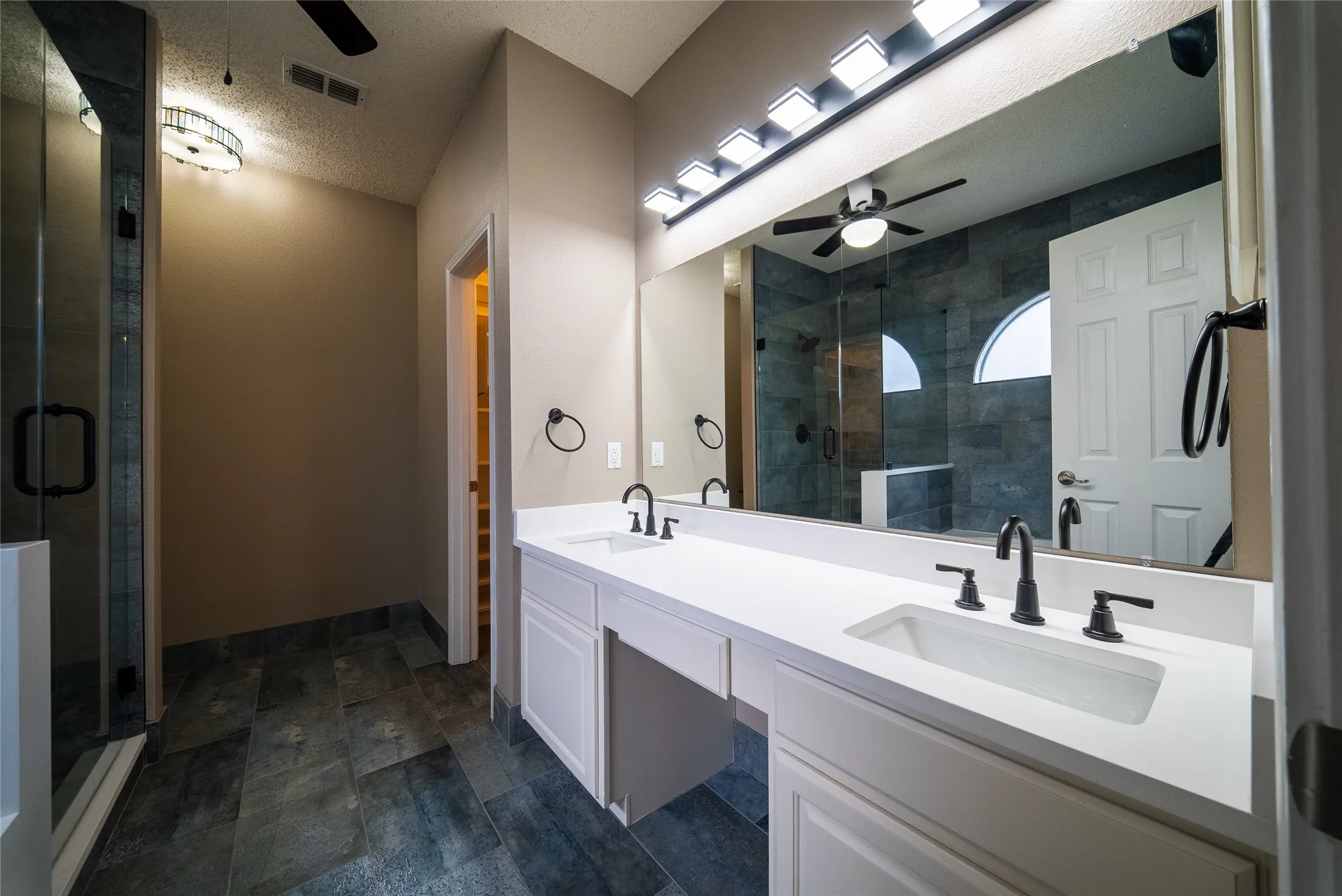 Dual-sink vanity with white stone-finish countertop, expansive wall-to-wall mirror, dark-toned tile flooring, and a glass-enclosed shower with tiled surround