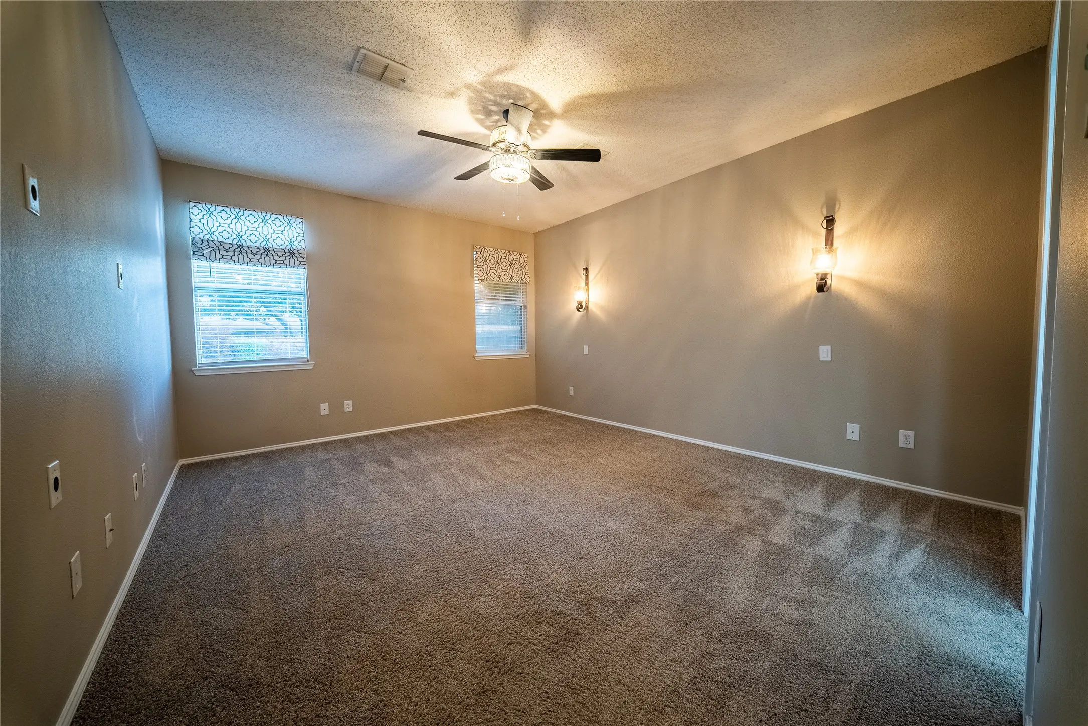 Neutral interior space featuring carpeted flooring, a ceiling fan with integrated lighting, dual windows, and wall-mounted sconce lighting