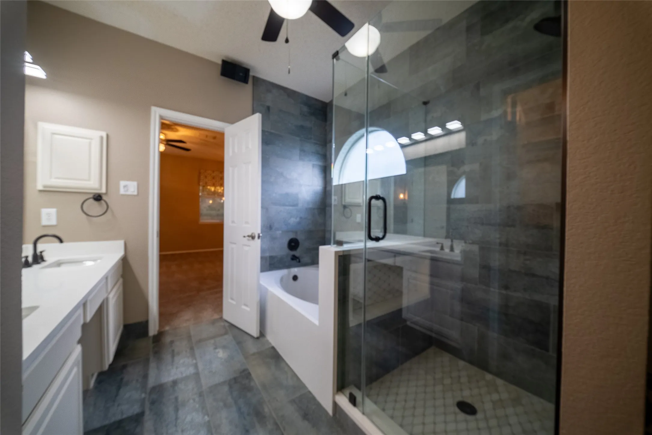 Bathroom featuring a walk-in shower with glass enclosure, soaking tub, slate-finish wall and floor tiling, and a dual-sink vanity with white countertop