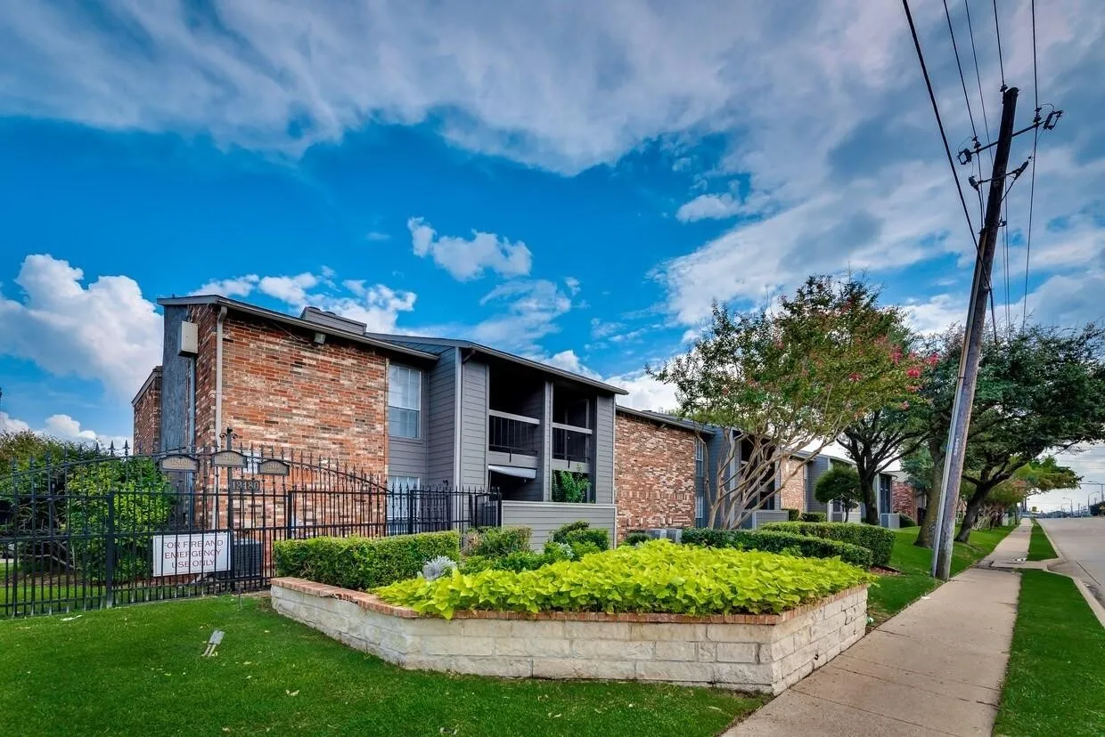 Multi-story building exterior featuring red brick and dark-toned paneling, raised stone-block landscaping, wrought-iron perimeter fencing, and mature trees