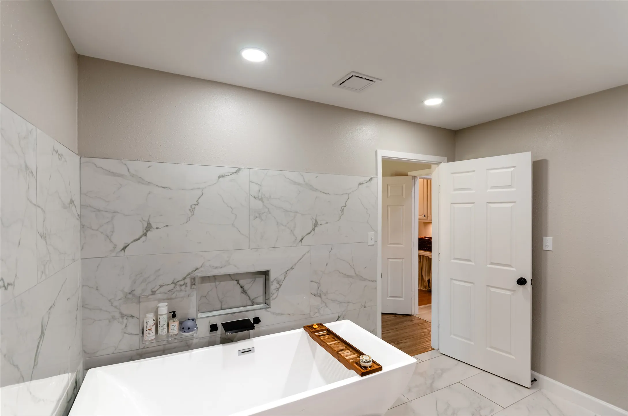 Freestanding soaking tub with a wall-mounted faucet, marble-look tile flooring, and matching marble-look wall tile with an integrated niche
