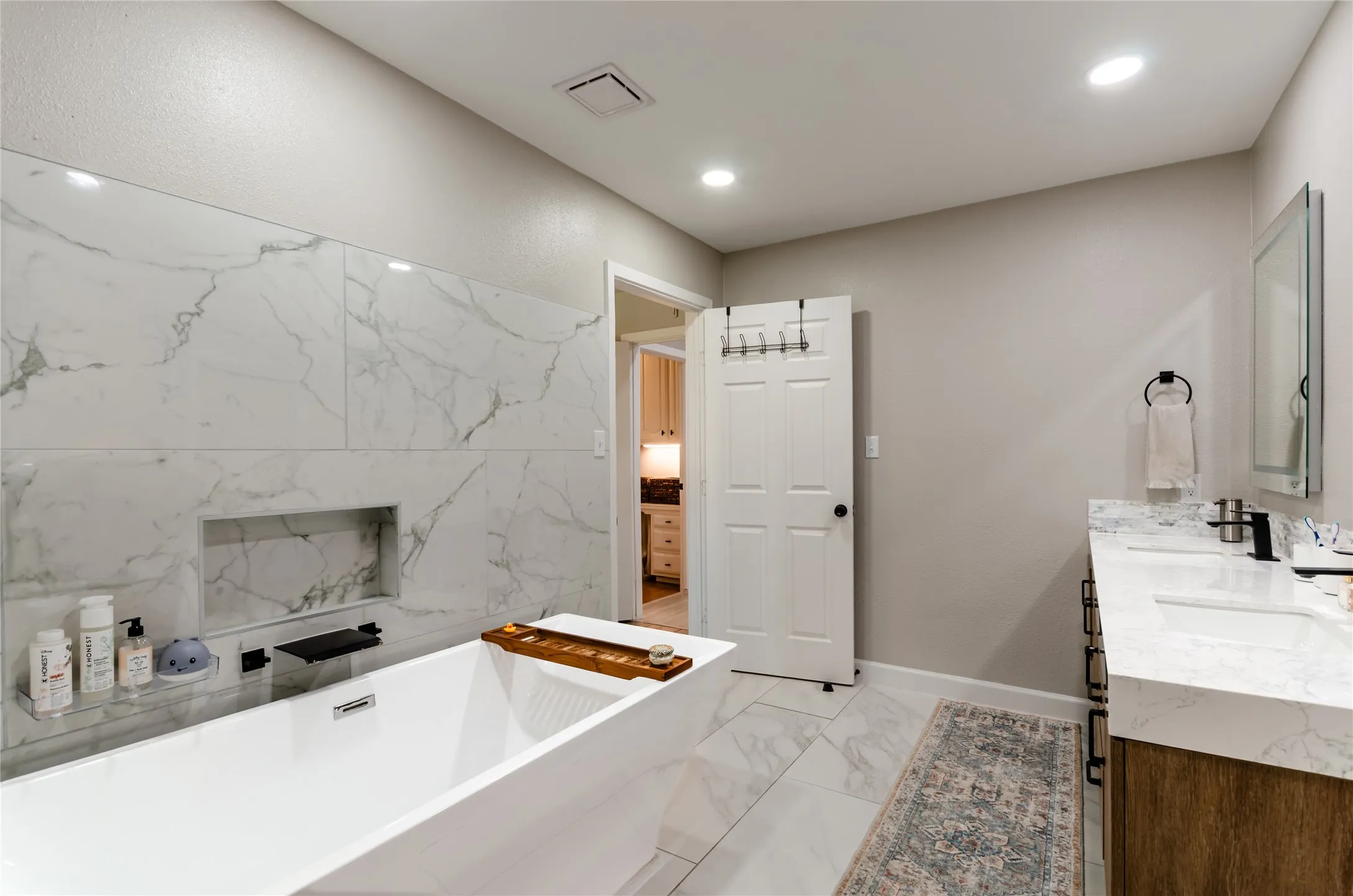 Freestanding soaking tub with a built-in wall niche, dual sink vanity with stone countertops, and recessed lighting
