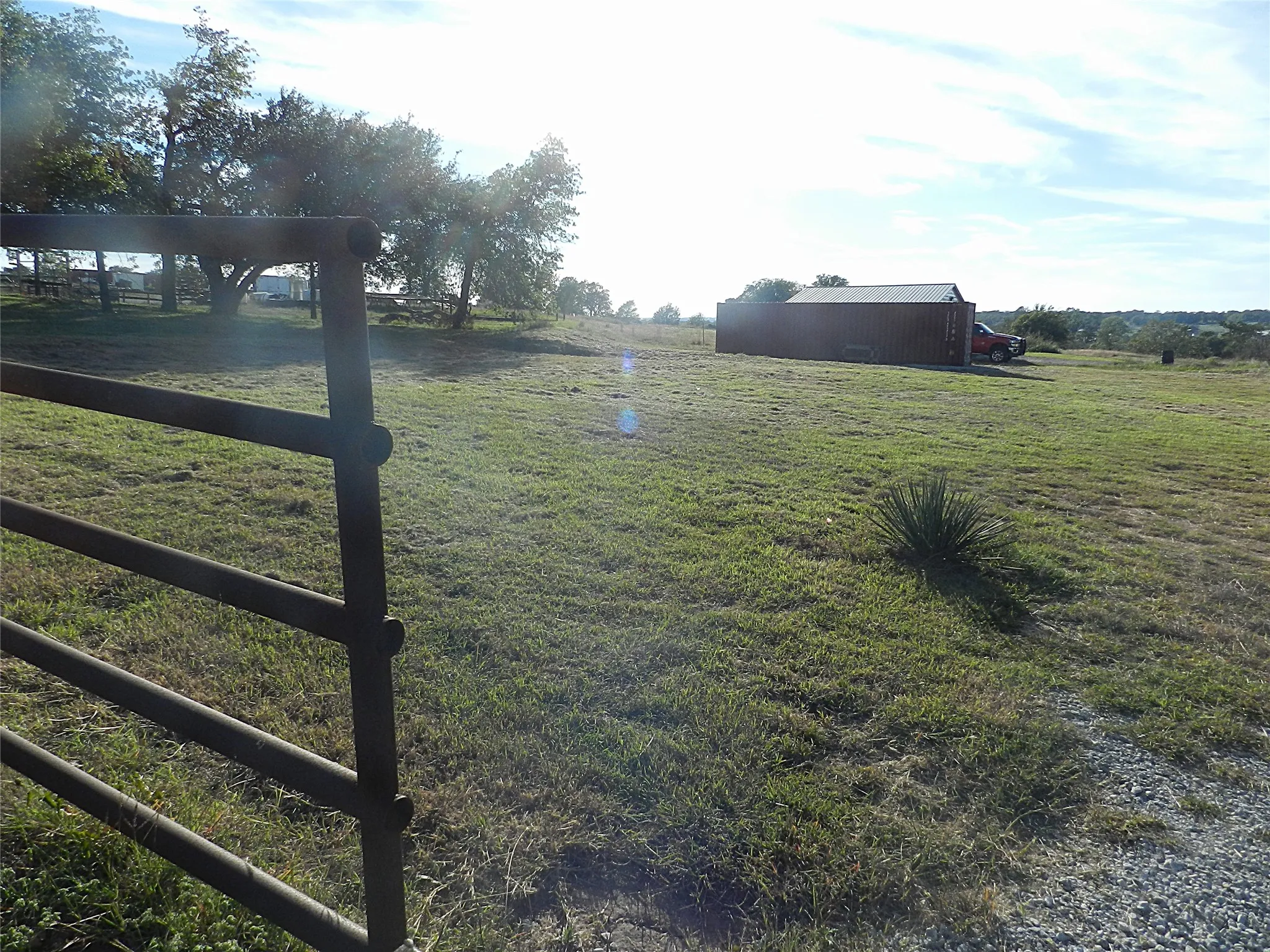 Open acreage featuring a metal tubular gate, expansive grass pasture, mature trees, and a detached metal storage structure