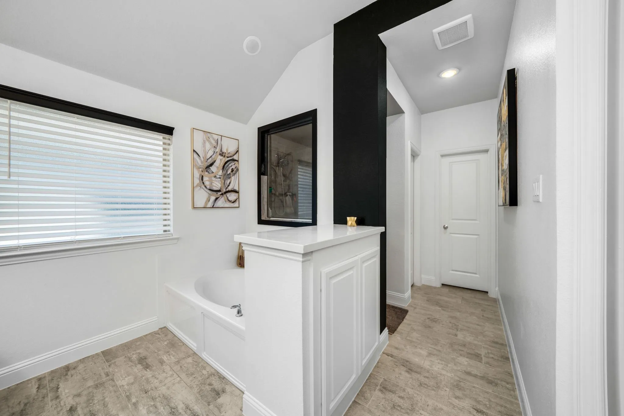 Soaking tub with white surround, stone-finish tile flooring, white shaker-style cabinetry with quartz countertop, high-contrast black architectural accent wall, and horizontal slat window blinds
