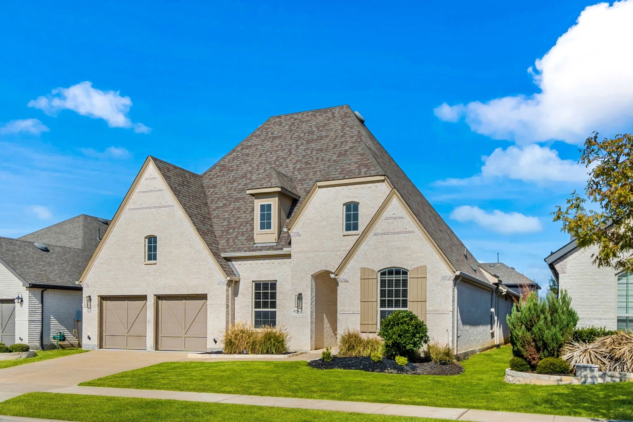 Two-story exterior featuring light-toned brick masonry, a complex multi-gabled roofline, decorative shutters, and a two-car garage with carriage-style doors