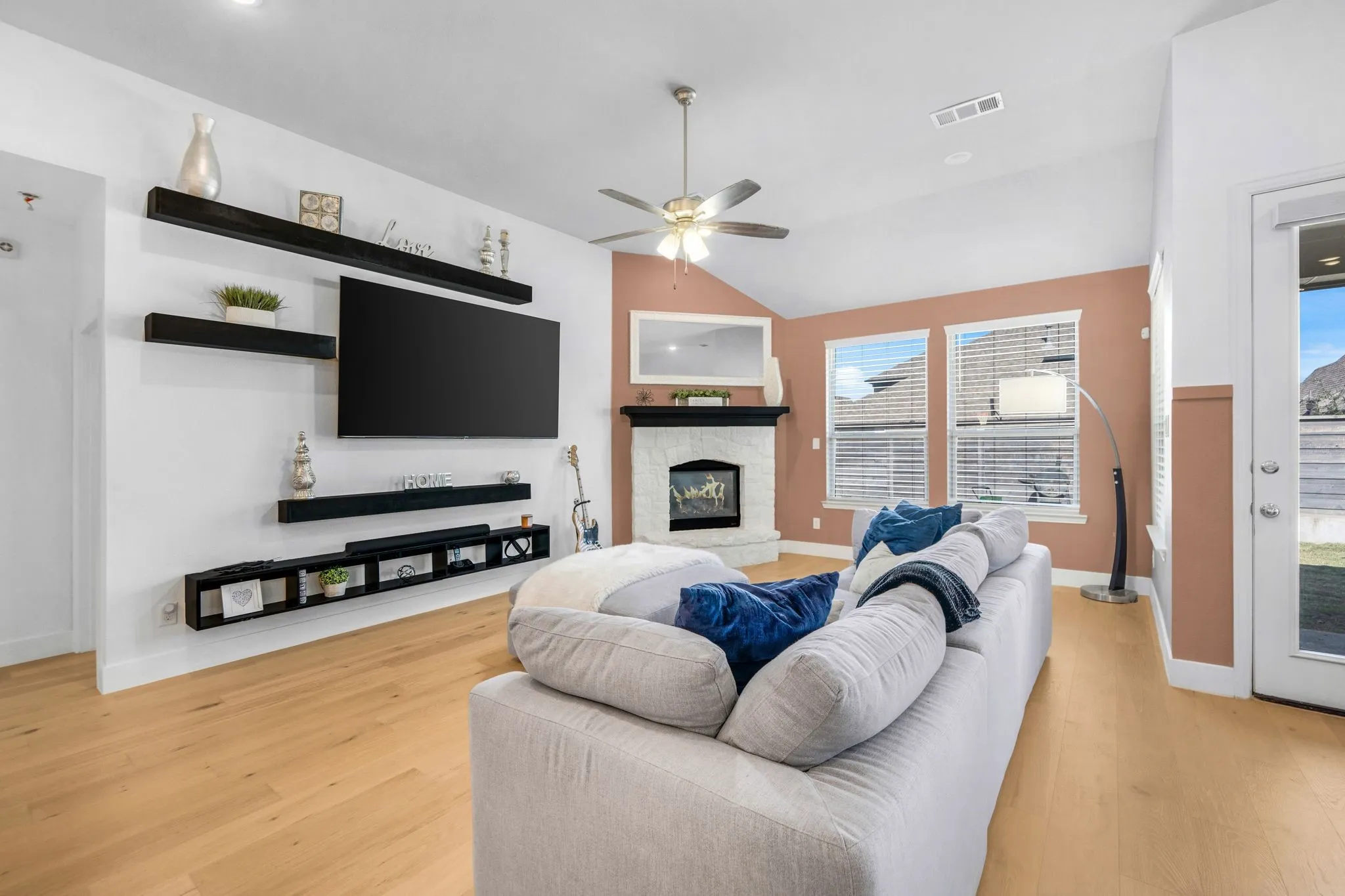 Vaulted ceiling with a central fan, wood-finish flooring, stone-surround fireplace with a mantel, and built-in wall shelving