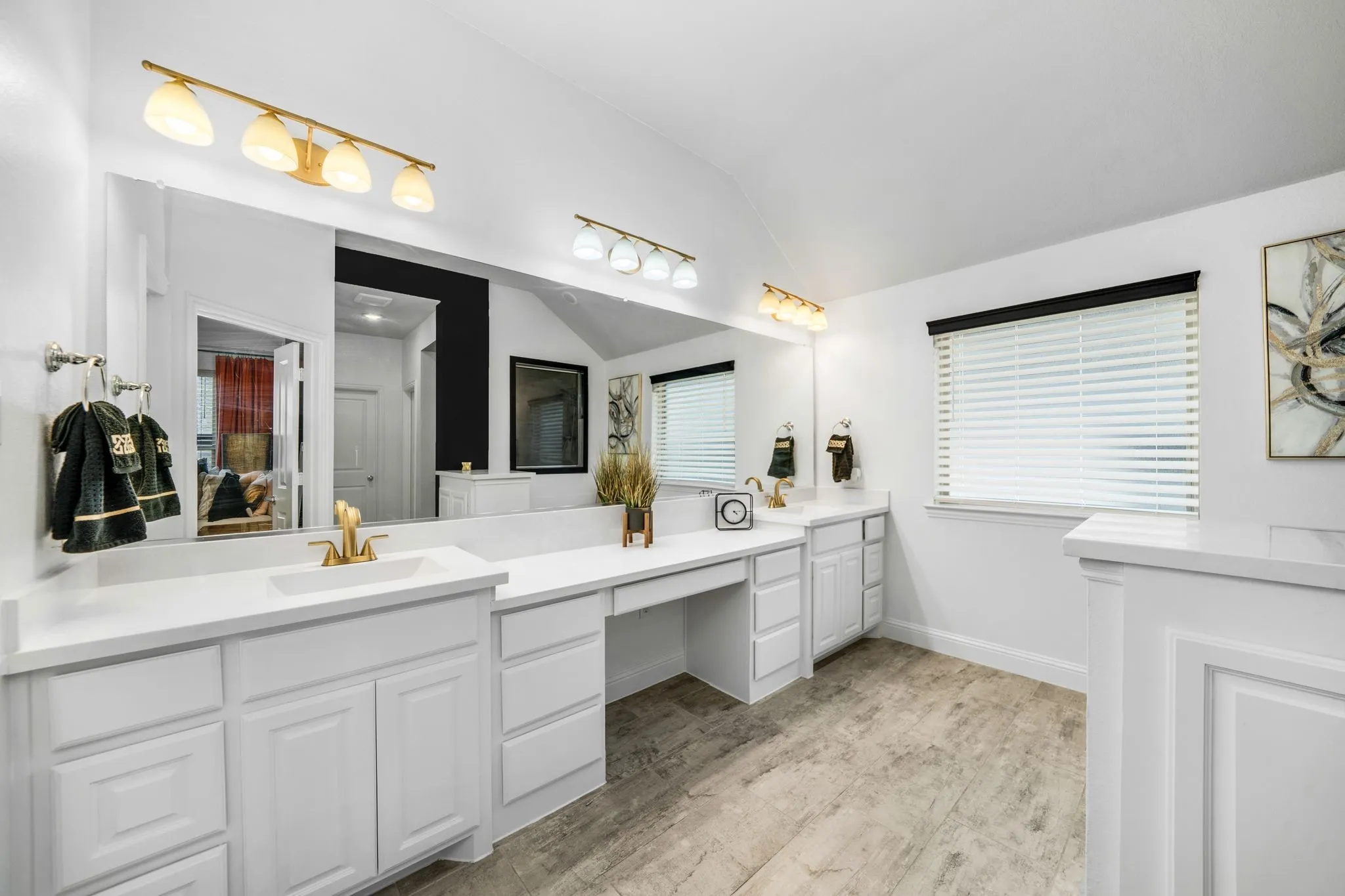 Dual-sink vanity with white cabinetry, integrated stone-surface countertop, wood-finish flooring, full-width vanity mirror, and three-piece multi-bulb lighting fixtures