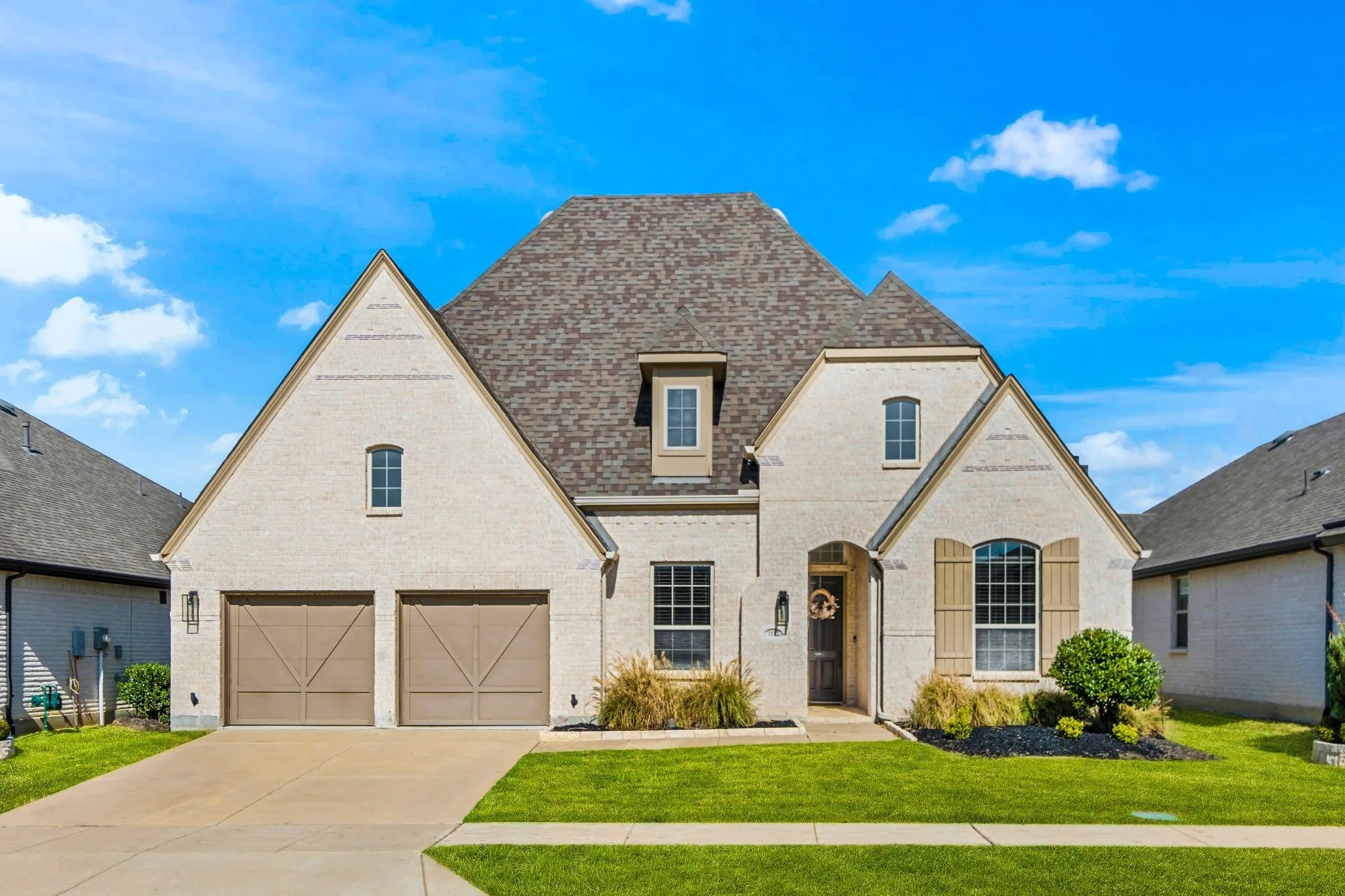 Multi-gabled exterior featuring light-toned brick masonry, a steeply pitched roof, arched entryway, wooden-finish garage doors, and decorative shutters