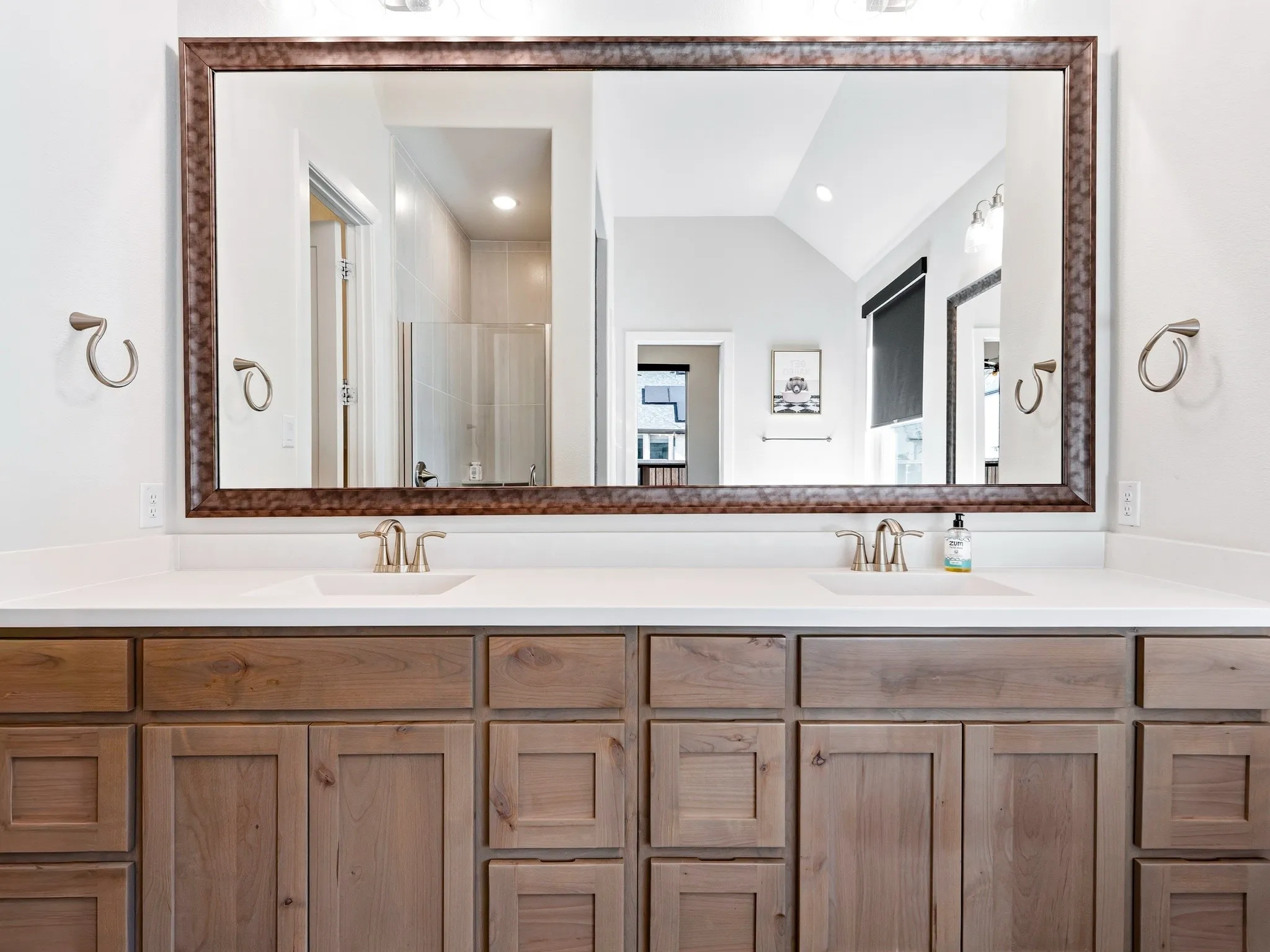 Dual-sink vanity with a solid-surface countertop, wood-finish shaker cabinetry, large framed mirror, and dual brushed-finish faucets