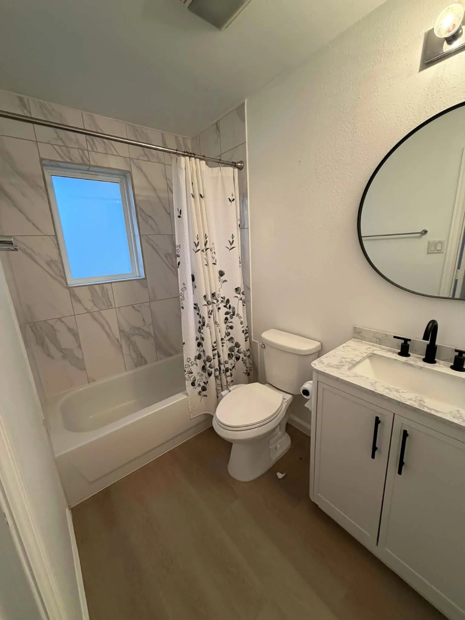 2nd Bathroom featuring a vanity with a stone-finish countertop, undermount sink, and matte black hardware, paired with a shower-tub combination featuring floor-to-ceiling stone-patterned wall tile and a circular vanity mirror