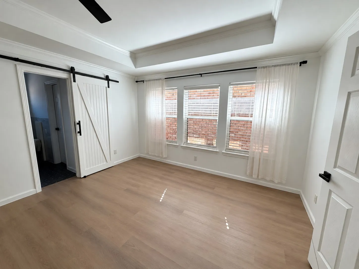 Tray ceiling with recessed lighting, wood-finish flooring, sliding white barn door with matte black hardware, and three-pane window configuration