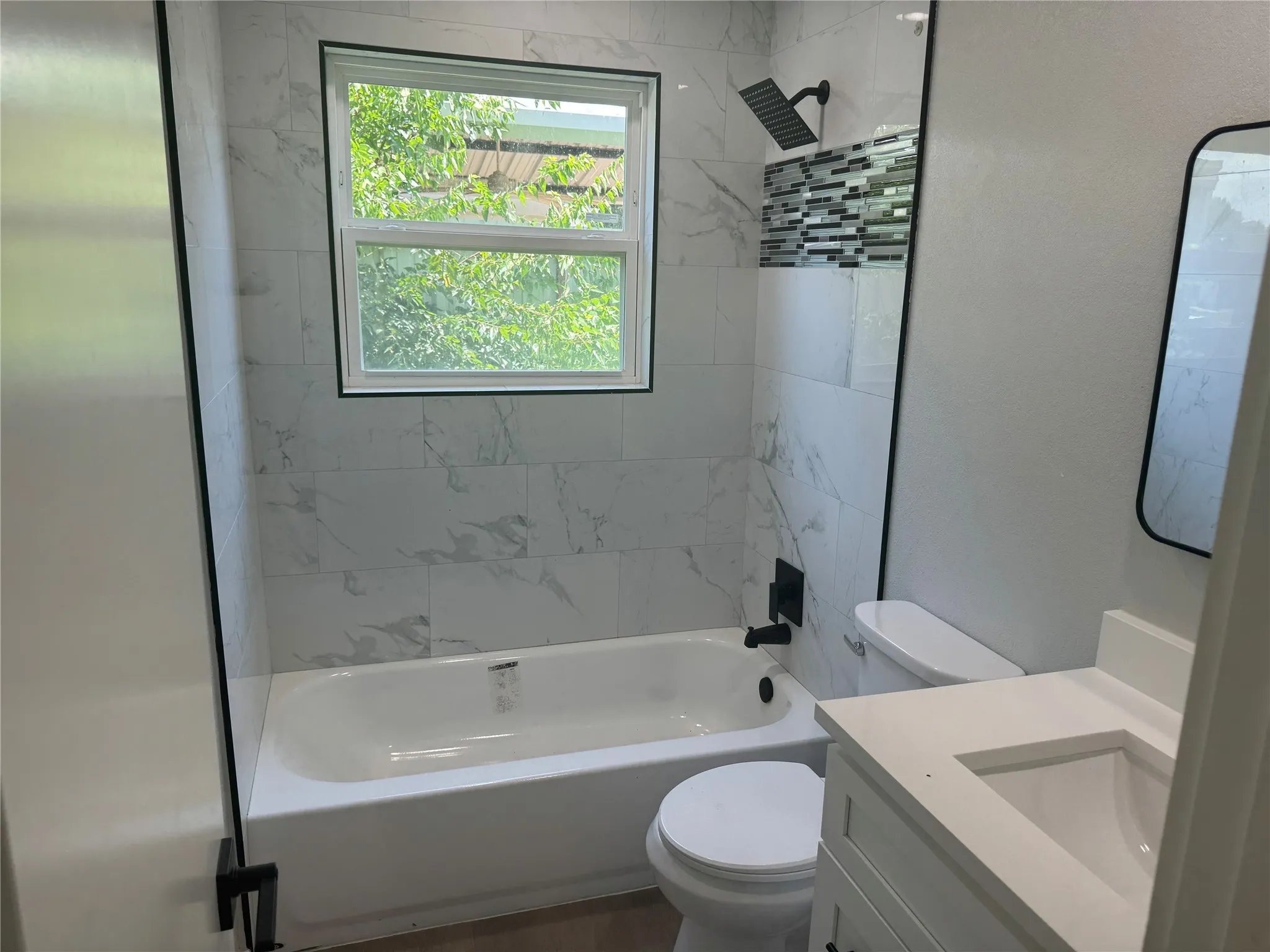 Bathroom featuring stone-finish wall tile, a built-in tub with matte black rain shower head and hardware, an integrated vanity, and a rectangular mirror
