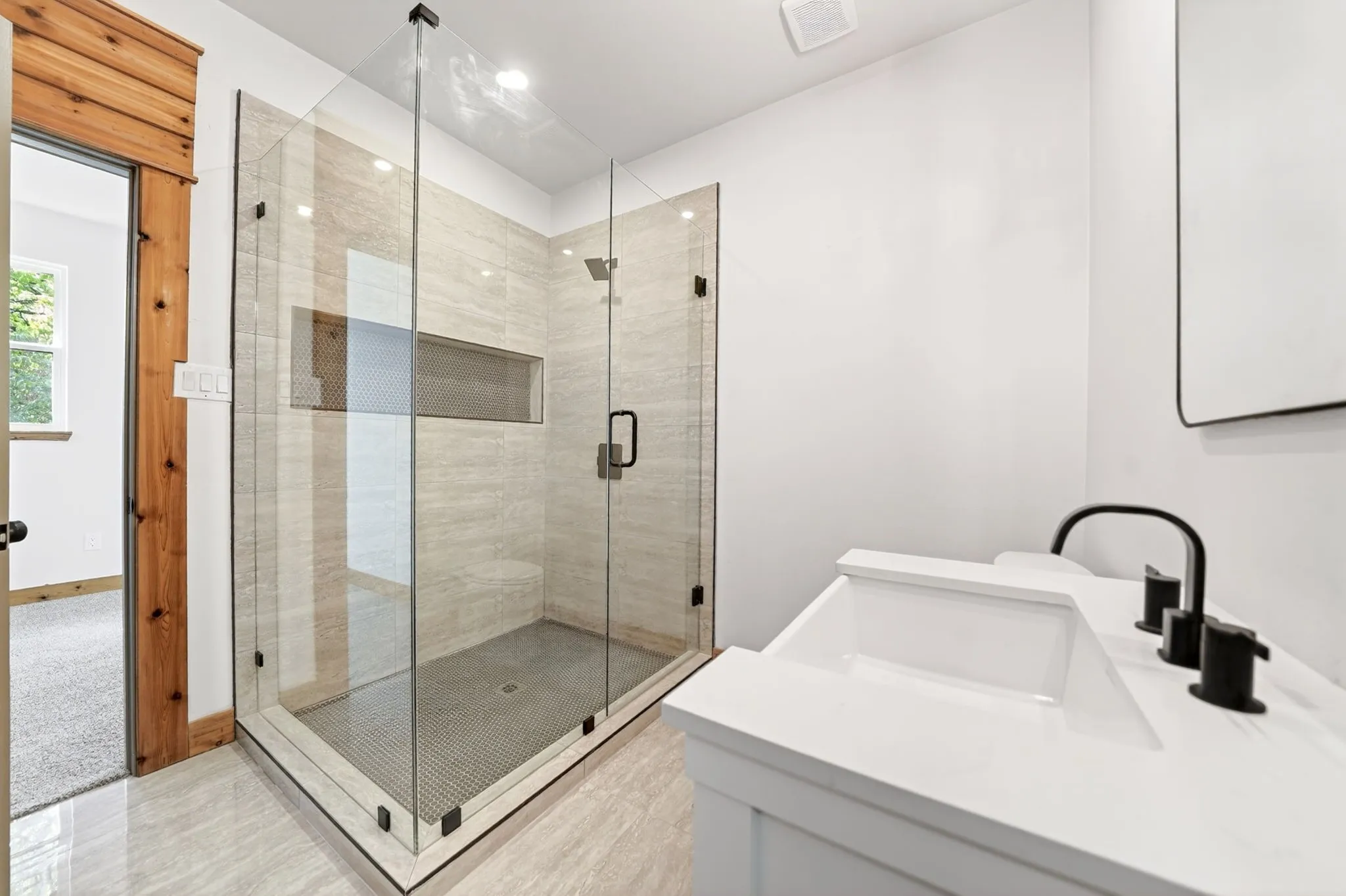 Frameless glass-enclosed shower with tiled walls and built-in niche, white vanity cabinet with stone-finish countertop, and matte black plumbing fixtures