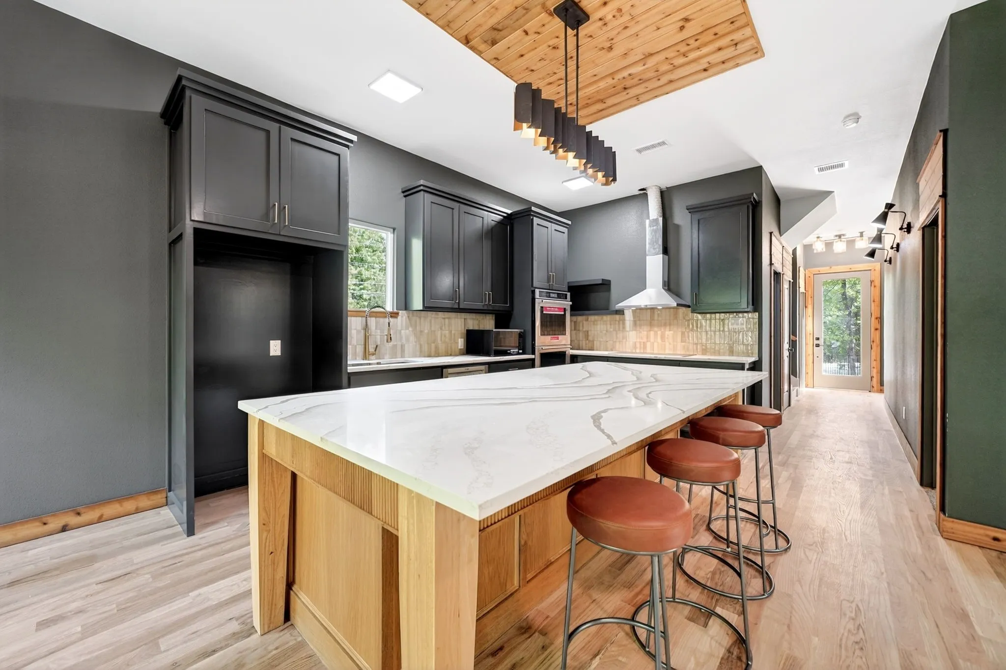 Kitchen featuring an expansive center island with a stone-surface countertop, ceiling-mounted wood inlay detail, charcoal-toned cabinetry, stainless steel wall-mounted range hood, and light-tone wood-finish flooring