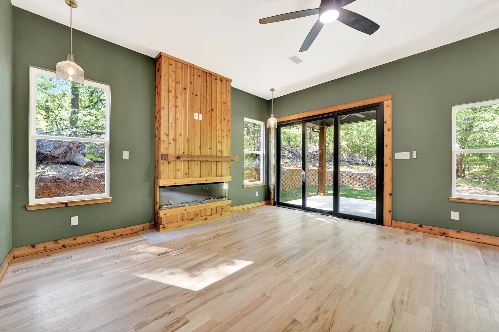 Living area featuring wood-finish flooring, a floor-to-ceiling wood-paneled fireplace, glass sliding doors, dual pendant lighting, and a ceiling fan