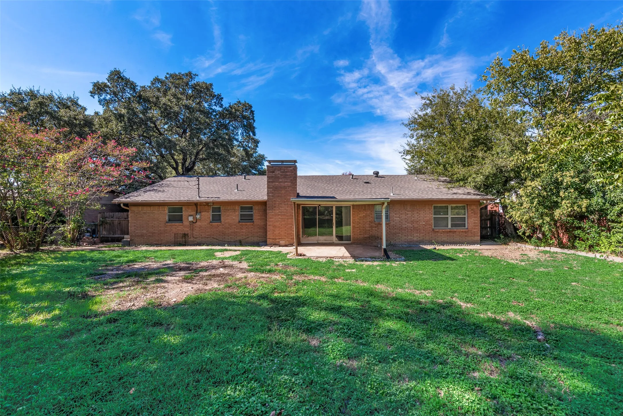 Rear elevation featuring full-brick masonry, a center brick chimney, concrete patio, and mature tree canopy