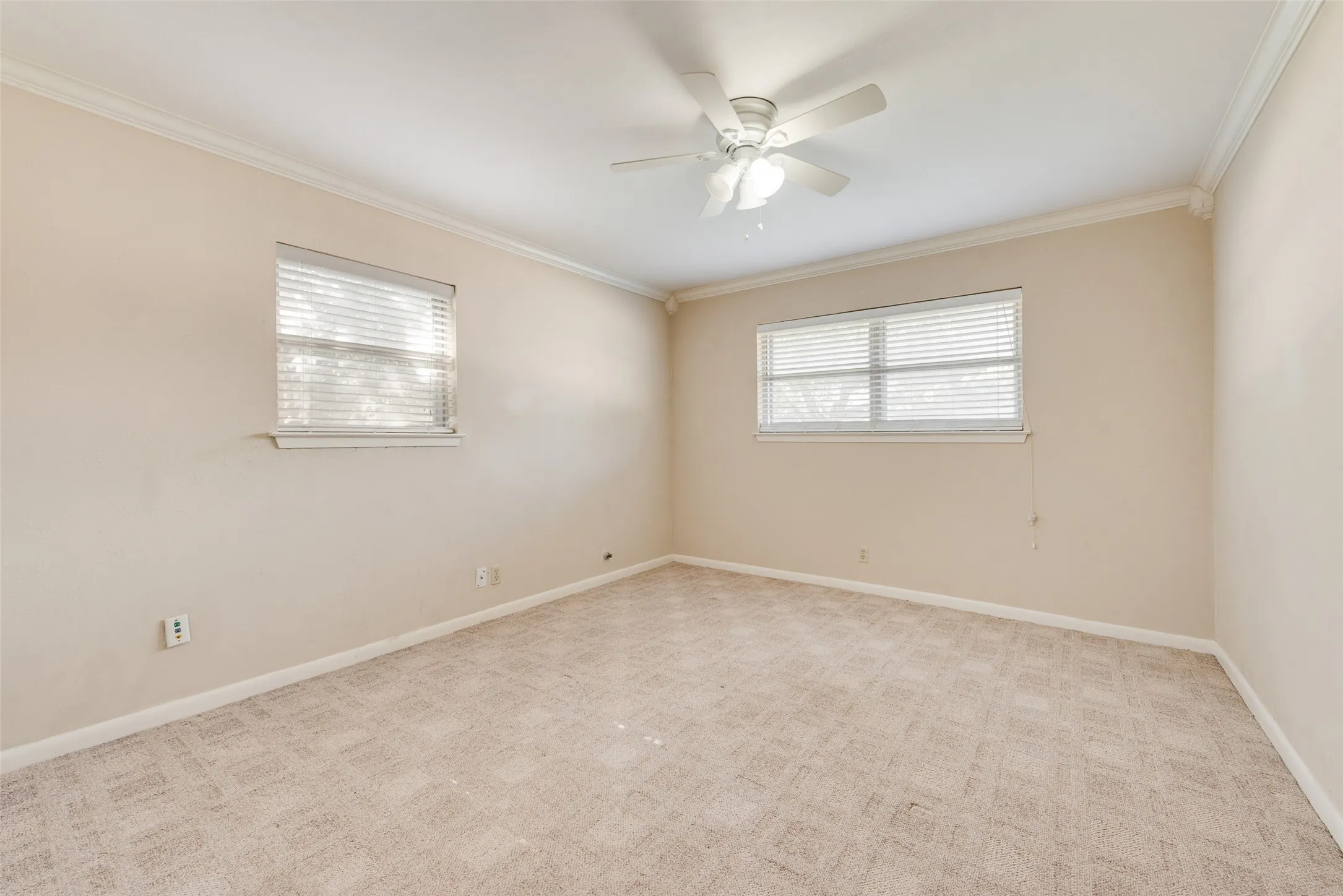 Neutral interior space featuring textured wall-to-wall carpeting, dual windows with integrated blinds, crown molding, and a central ceiling fan with integrated lighting