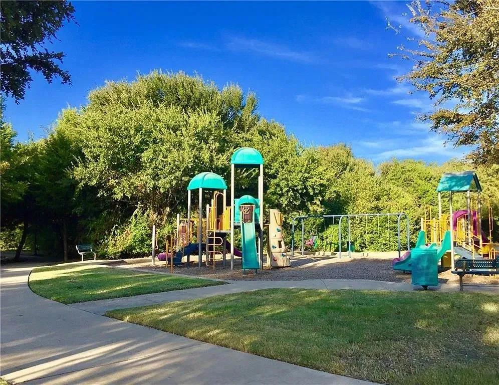 Outdoor recreation area featuring a multi-tiered playground structure, paved concrete walkways, benches, and manicured lawn surroundings