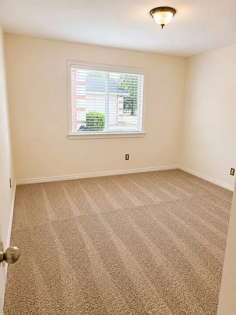 Neutral-toned interior space featuring textured wall-to-wall carpeting, a single-pane window with horizontal blinds, and a semi-flush mount ceiling fixture