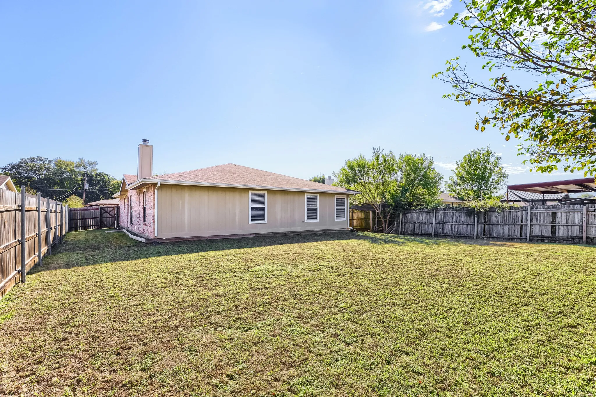Expansive backyard featuring a grass lawn, full-perimeter wood fencing, a masonry chimney, and a metal-roofed carport structure