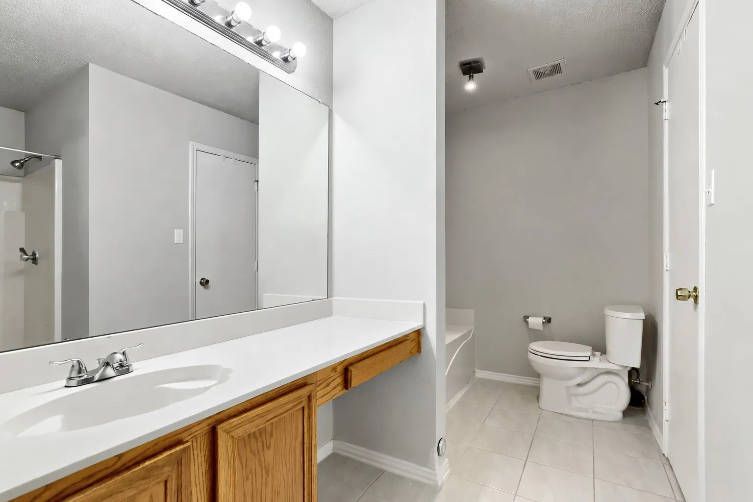 Full-width vanity with integrated sink, wood-finish cabinetry, expansive wall-mounted mirror, ceramic tile flooring, and separate water closet area
