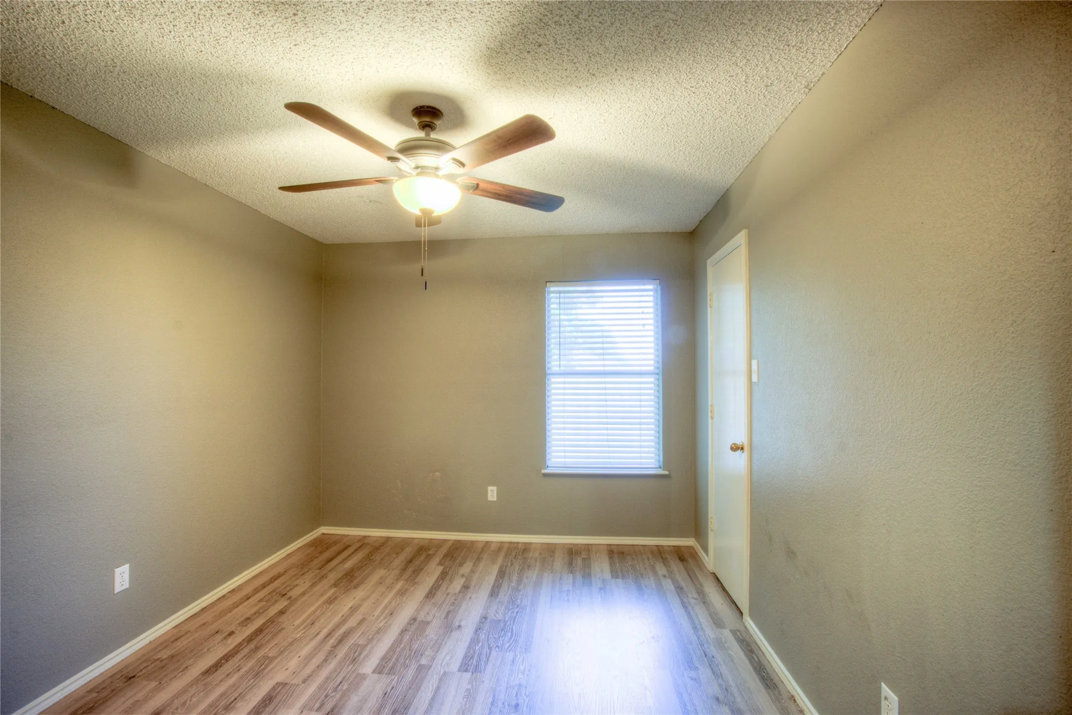 Room featuring wood-finish flooring, a ceiling fan with integrated lighting, a window with horizontal blinds, and a standard interior door