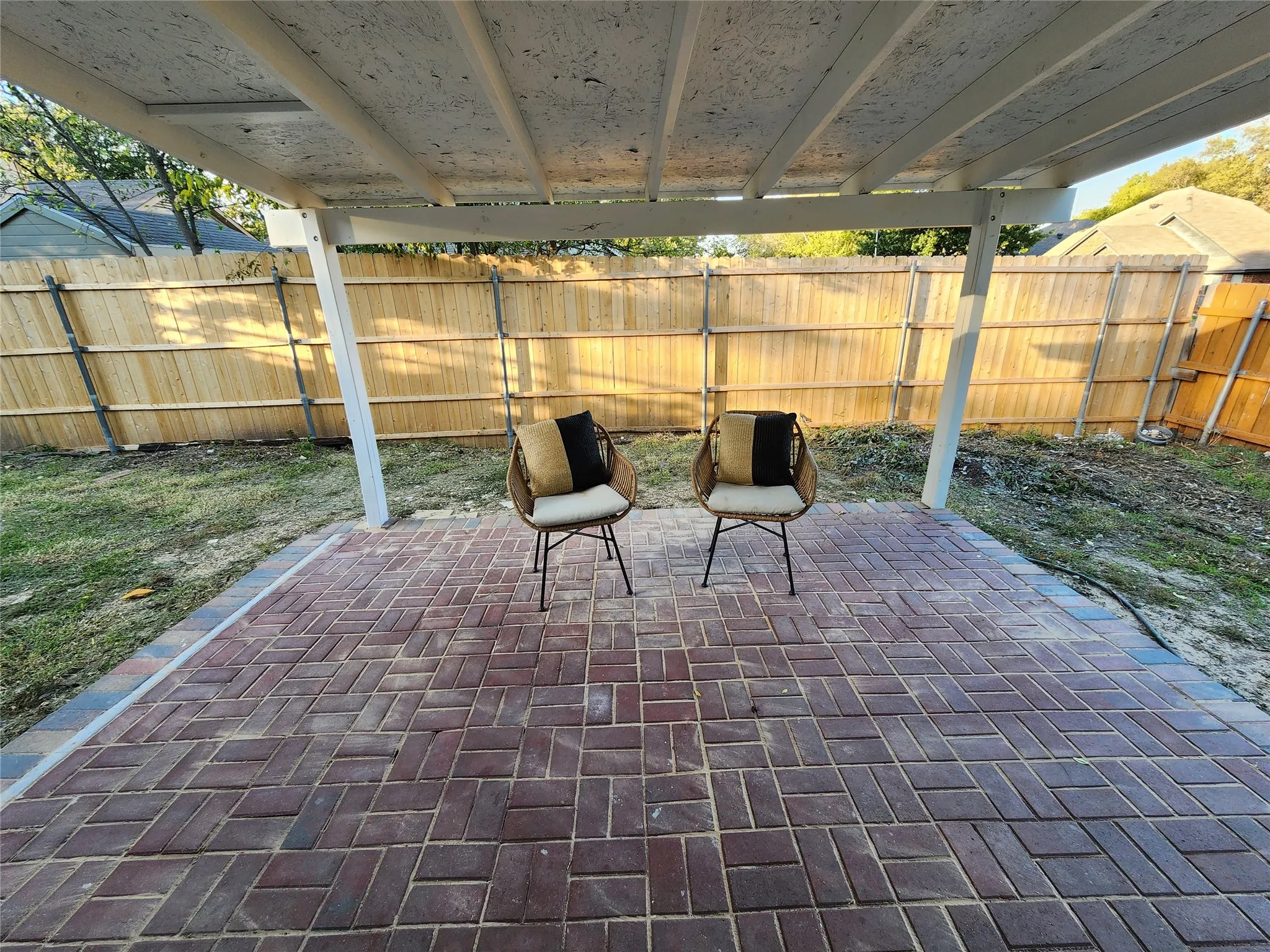 Covered patio featuring a herringbone-patterned brick paver floor and a structural wood-post frame, enclosed by a perimeter wood fence