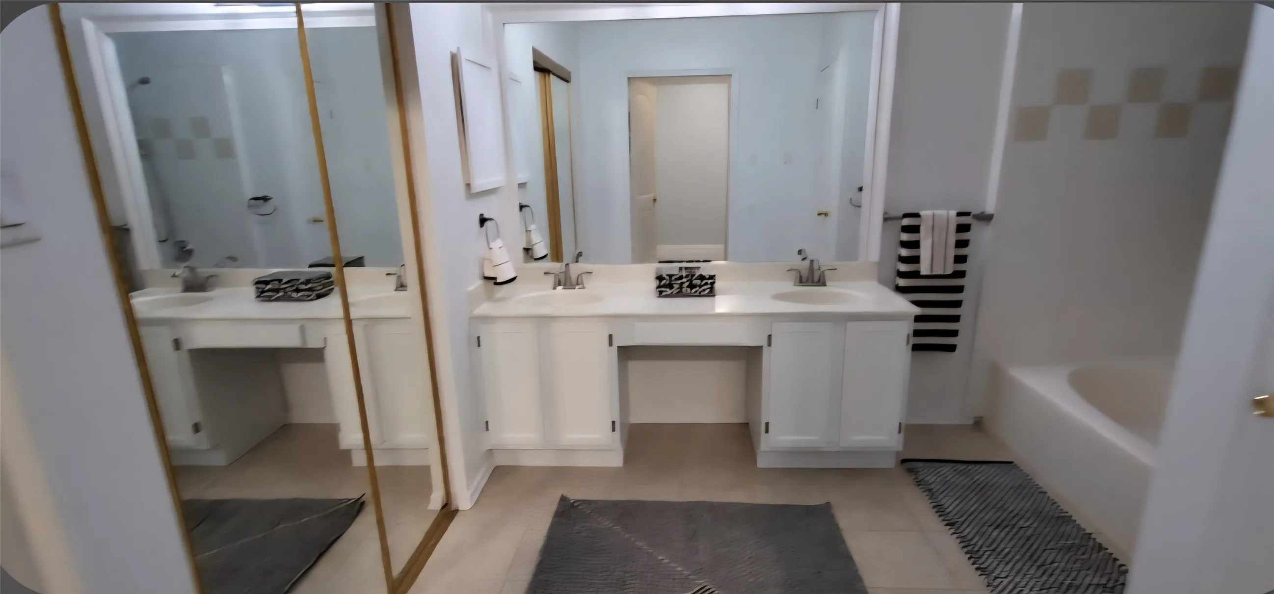 Primary Ensuite - Dual-sink vanity with white cabinetry, expansive wall-to-wall mirror, tiled tub surround, and mirrored wardrobe doors