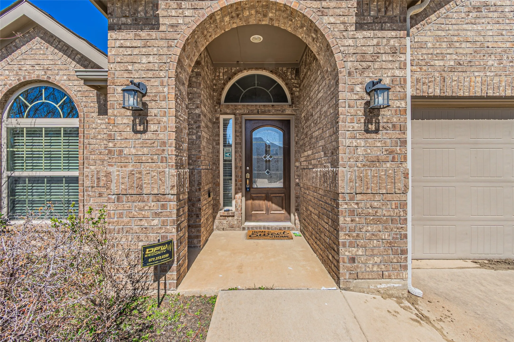 Brick facade entryway featuring an arched masonry portico, wood-finish front door with an integrated glass panel, transom window, and dual wall-mounted exterior lanterns