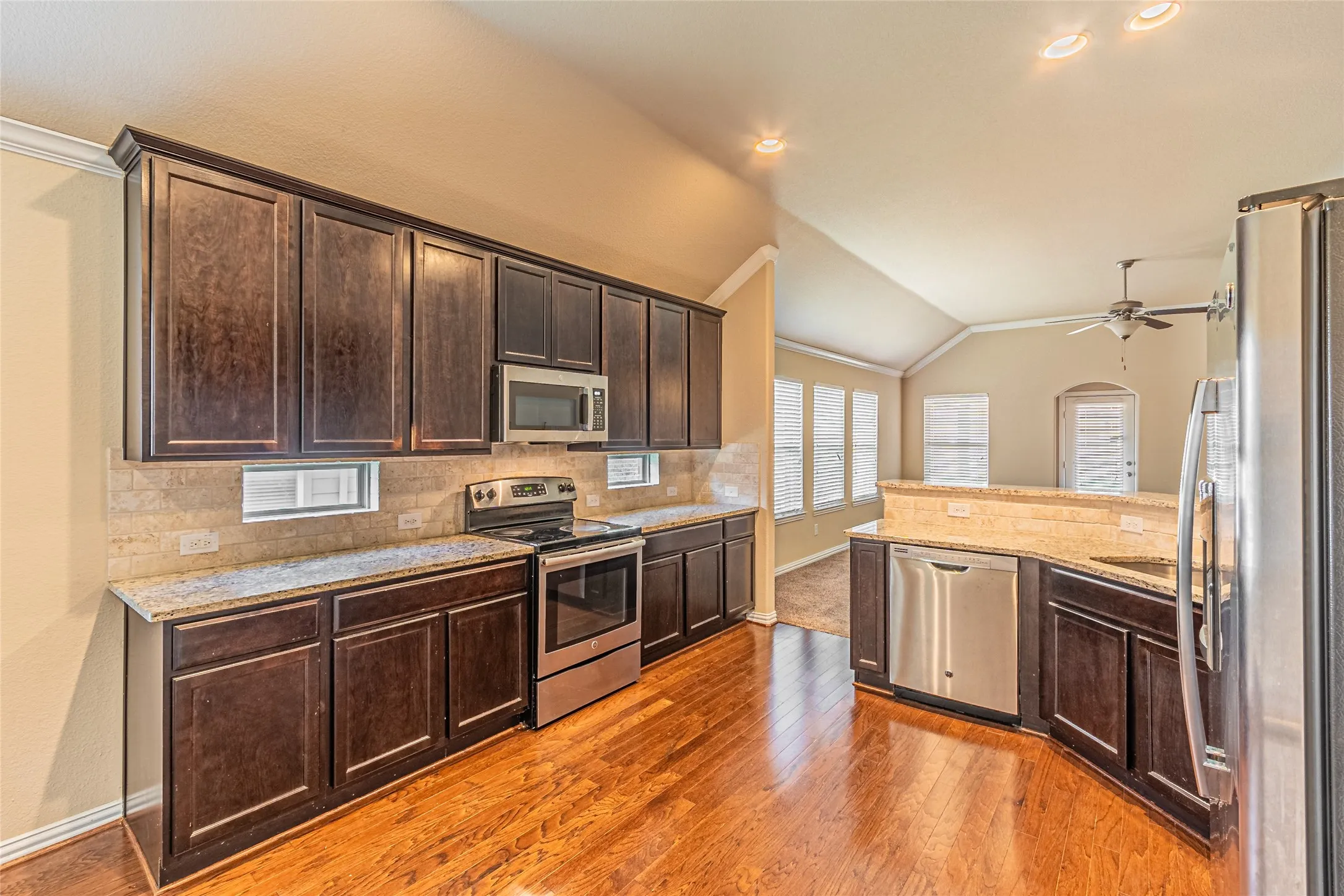 Kitchen featuring dark stained wood cabinetry, stone countertops, stainless steel appliances, tiled backsplash, and wood-finish flooring under vaulted ceilings