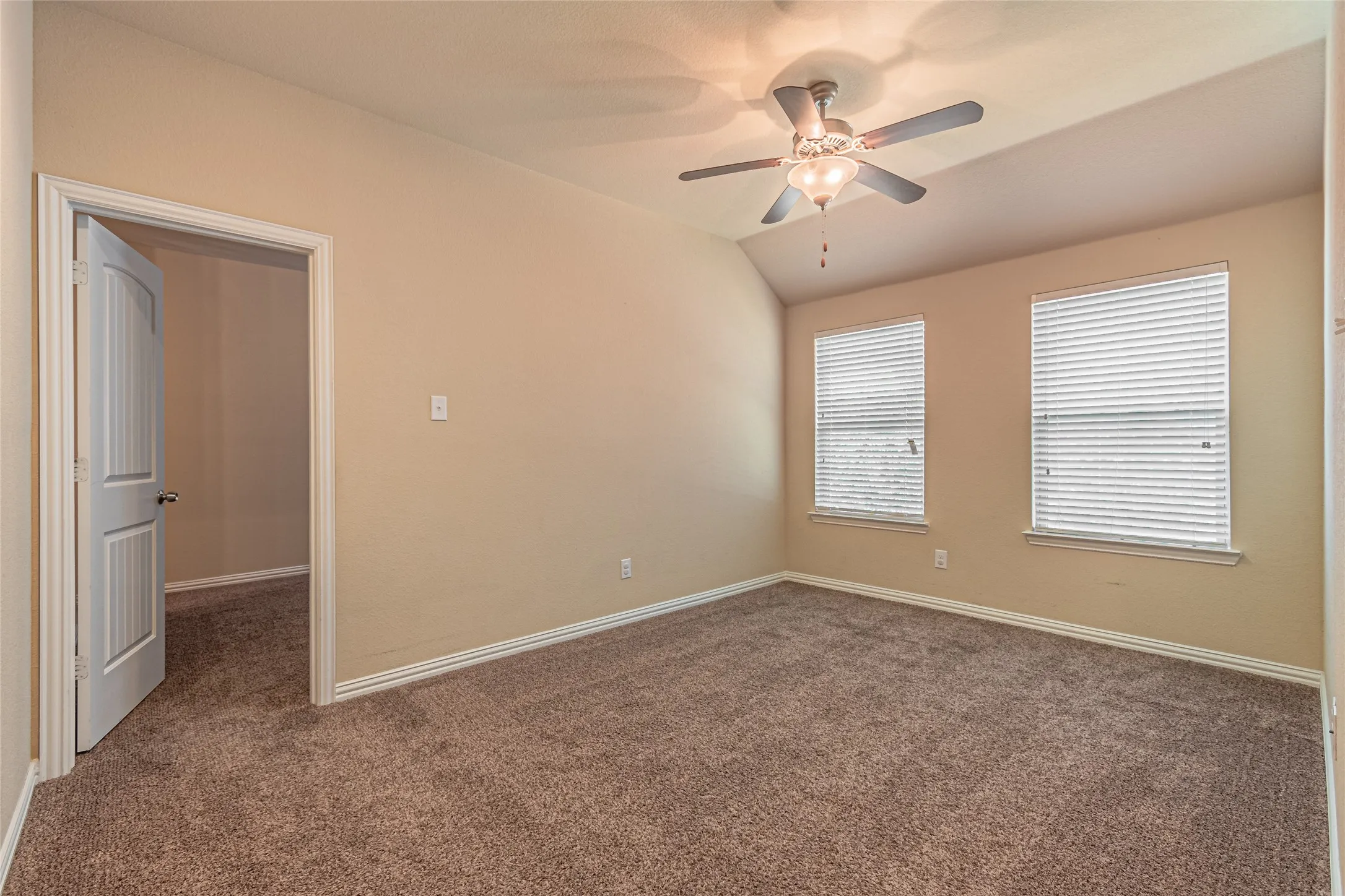 Room featuring neutral wall tones, plush wall-to-wall carpeting, a vaulted ceiling, dual windows with horizontal blinds, and a multi-blade ceiling fan