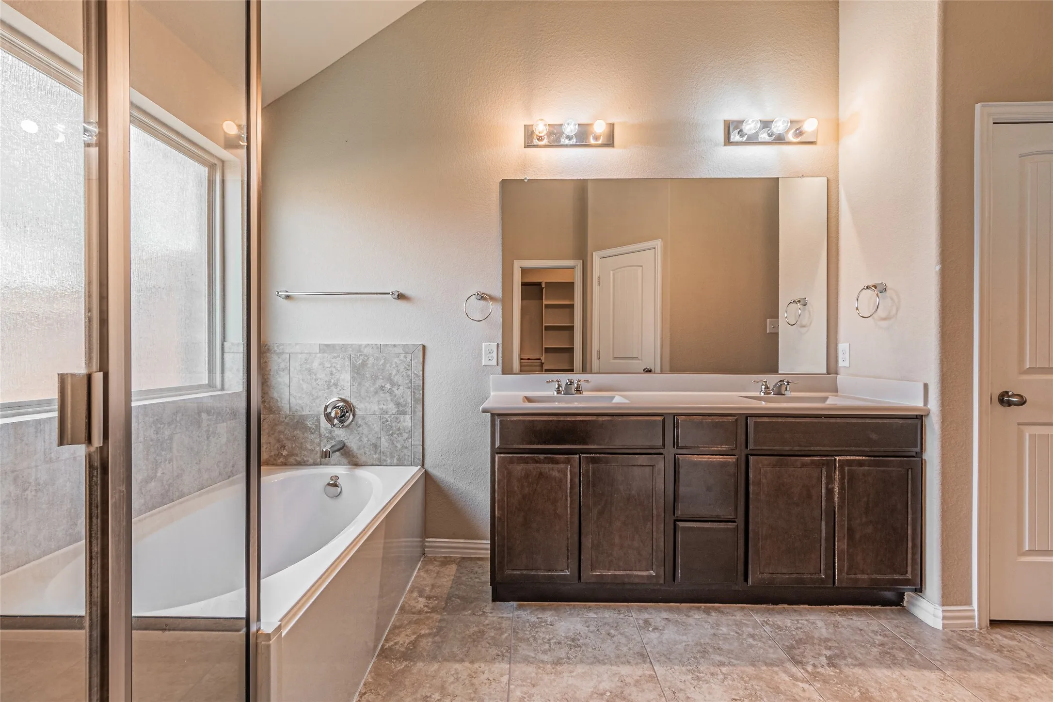 Bathroom featuring a dual-sink vanity with dark-finish cabinetry, a large wall-mounted mirror, a separate soaking tub with stone-textured surround, and a glass-enclosed shower