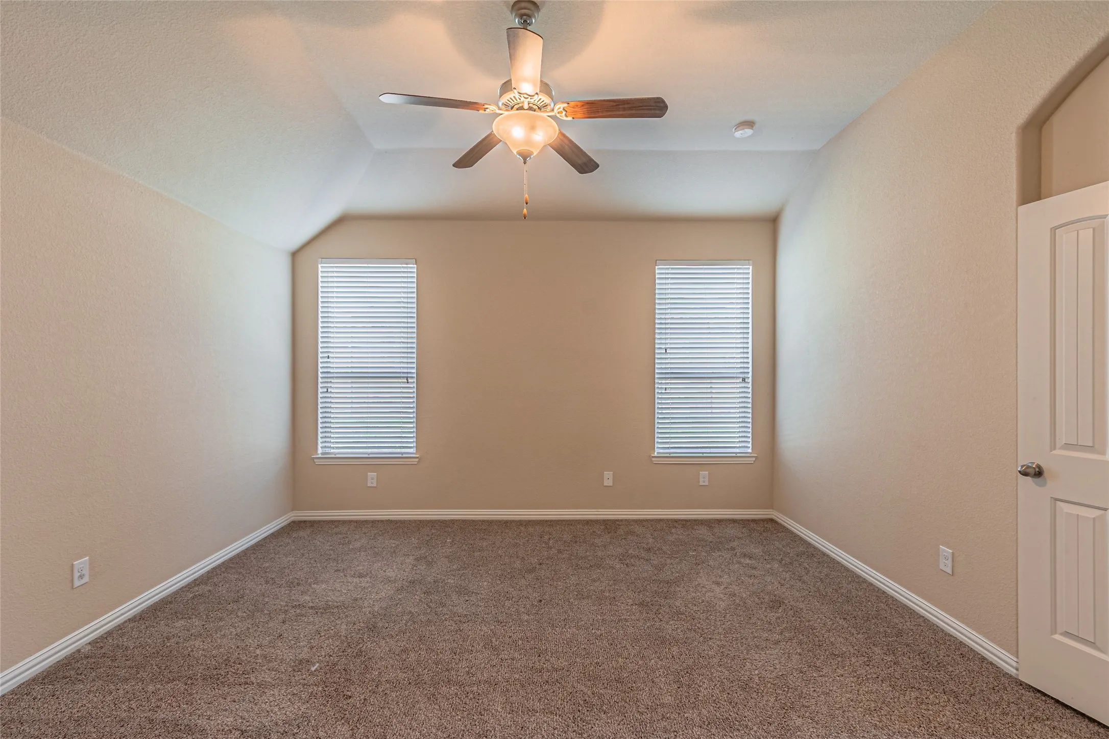 Room featuring a vaulted ceiling, wall-to-wall carpeting, two windows with horizontal blinds, and a centrally mounted ceiling fan with integrated light fixture