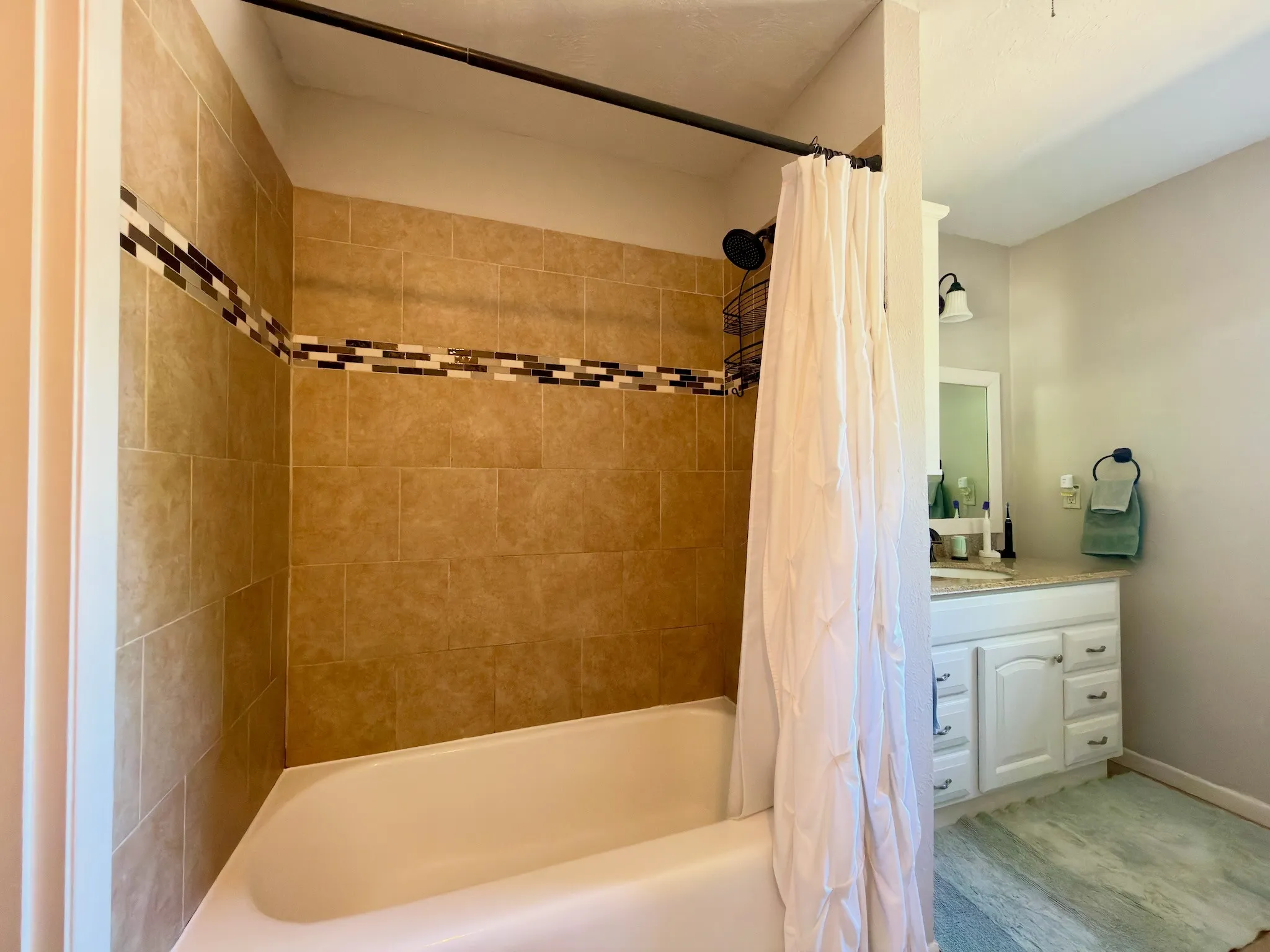 Bathroom featuring a tub with a tan tiled surround, decorative stone-finish tile accent band, built-in white cabinetry, stone-finish vanity countertop, and dark-finish hardware