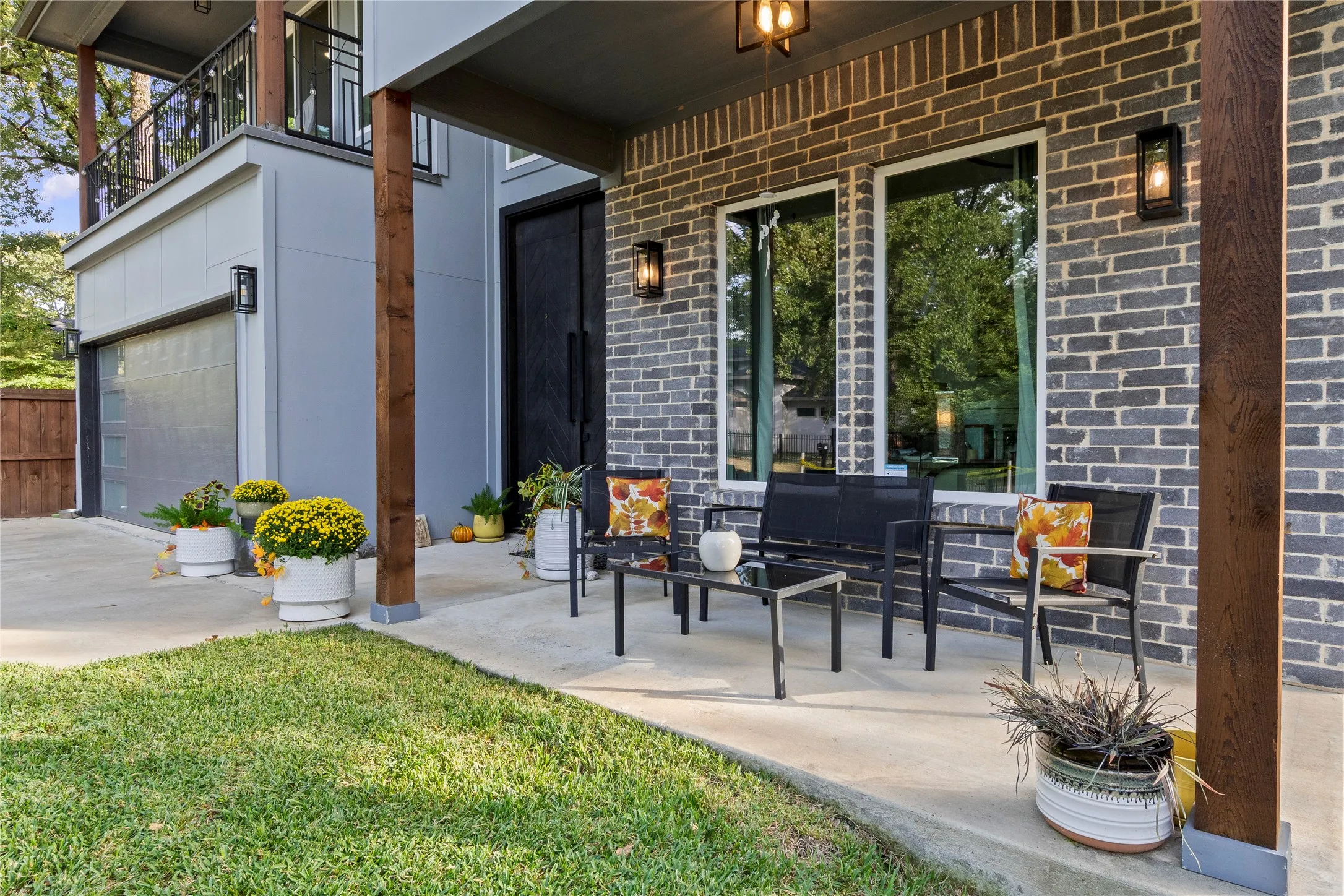 Covered patio featuring a grey brick facade, exposed wood-finish posts, double front doors, large windows, and wall-mounted exterior lighting