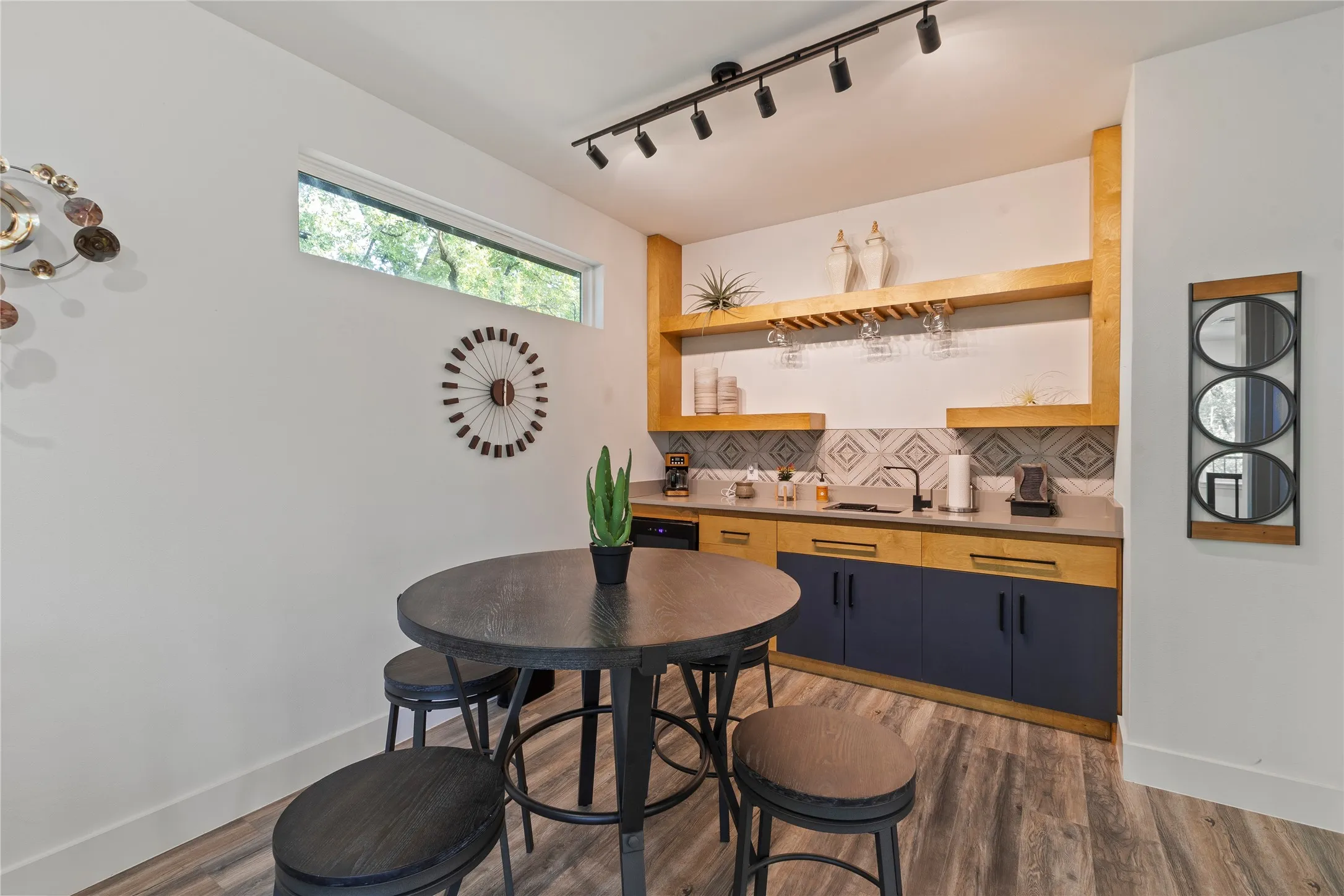 Built-in bar featuring custom dual-tone cabinetry, natural wood floating shelving, stone-finish countertops, geometric tile backsplash, and overhead track lighting