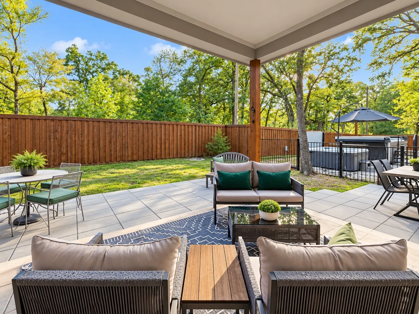 Covered patio featuring large-format stone pavers, wood-finished support column, privacy wood fencing, black metal perimeter fencing, and an integrated spa