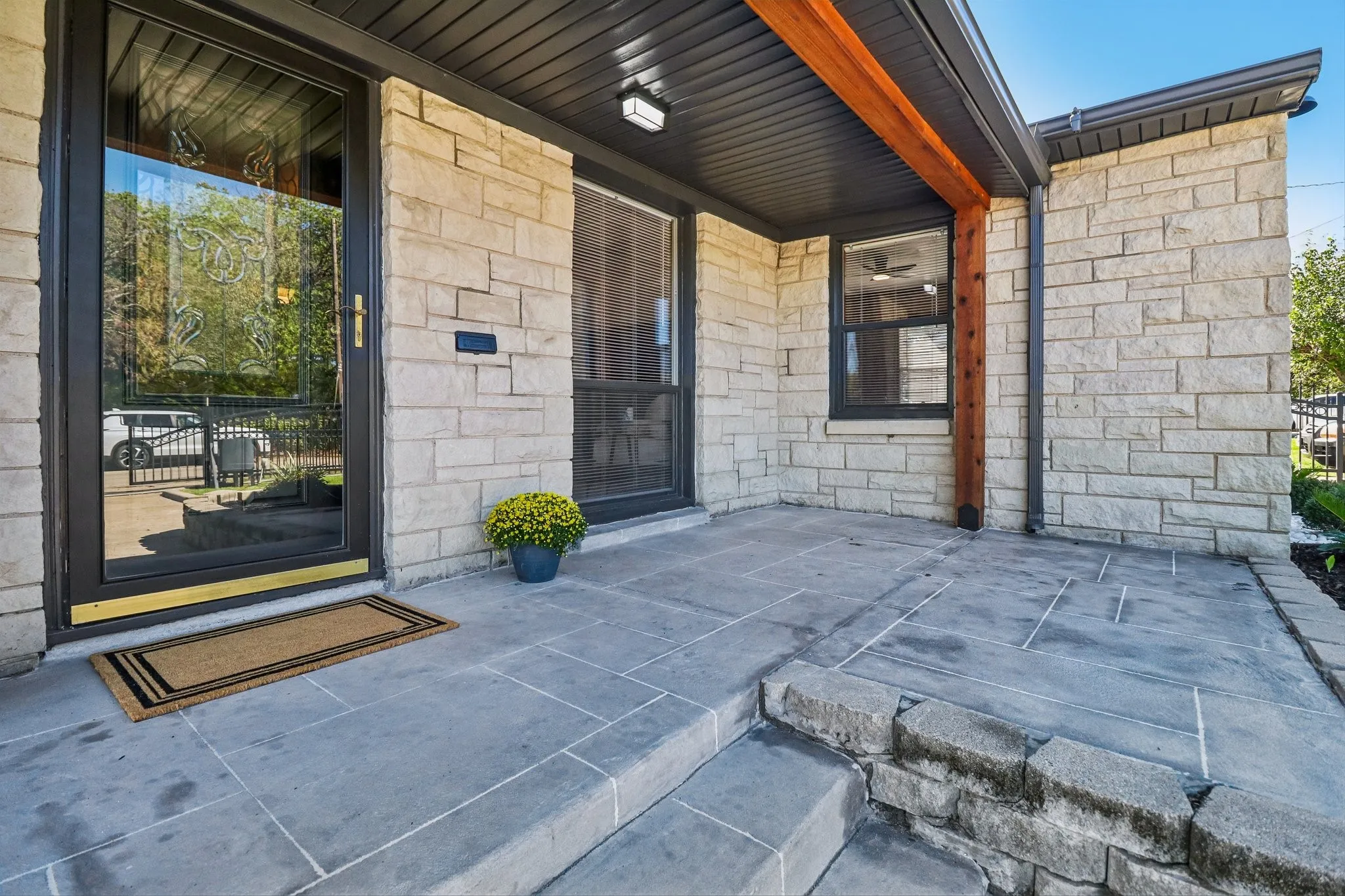 Stone veneer facade, glass-paneled entry door, dark-framed windows, wood beam accents, and a stone patio with integrated steps
