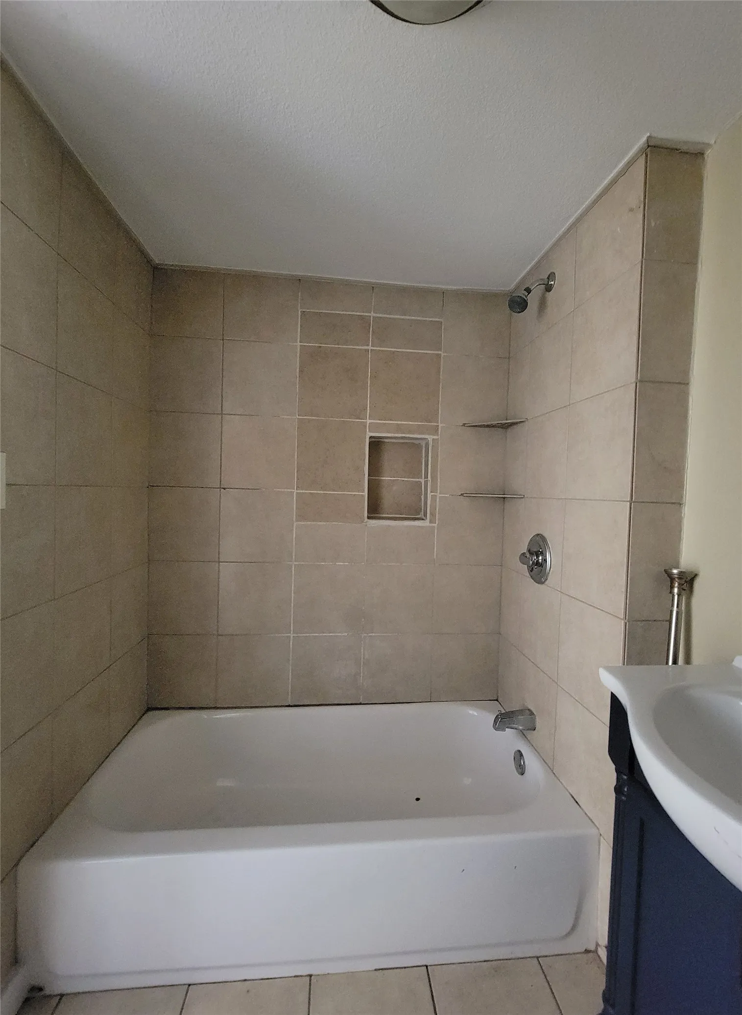 Bathtub with a shower head, tiled surround, built-in recessed wall niche, glass corner shelving, and metal plumbing fixtures