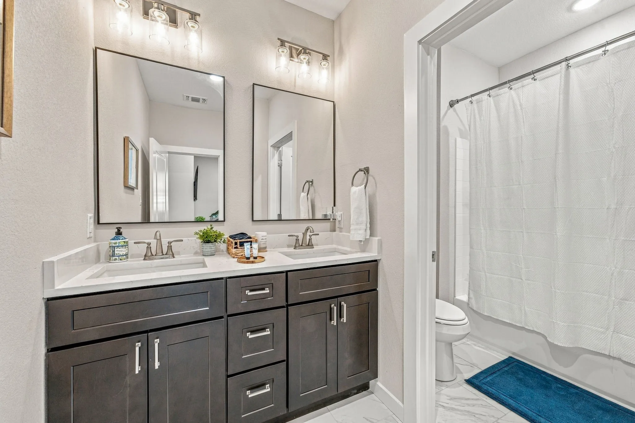 Dual-sink vanity featuring dark-toned cabinetry, light-colored stone-finish countertops, dual rectangular undermount sinks, and twin rectangular mirrors illuminated by multi-light wall-mounted fixtures